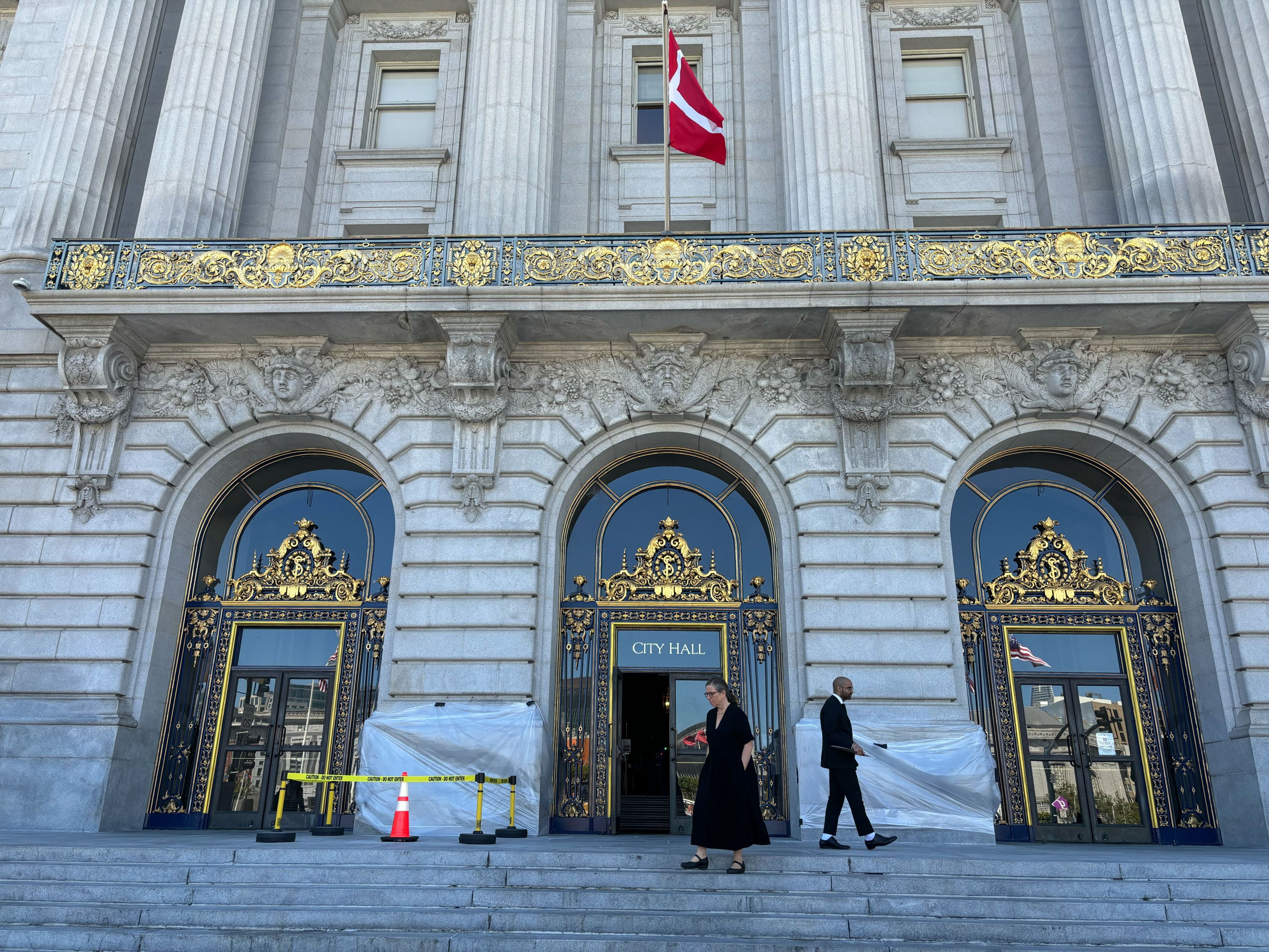 The image shows an ornate building facade with tall columns and gold detailing on arched doors. &quot;City Hall&quot; is written above the center door. Two people walk past.