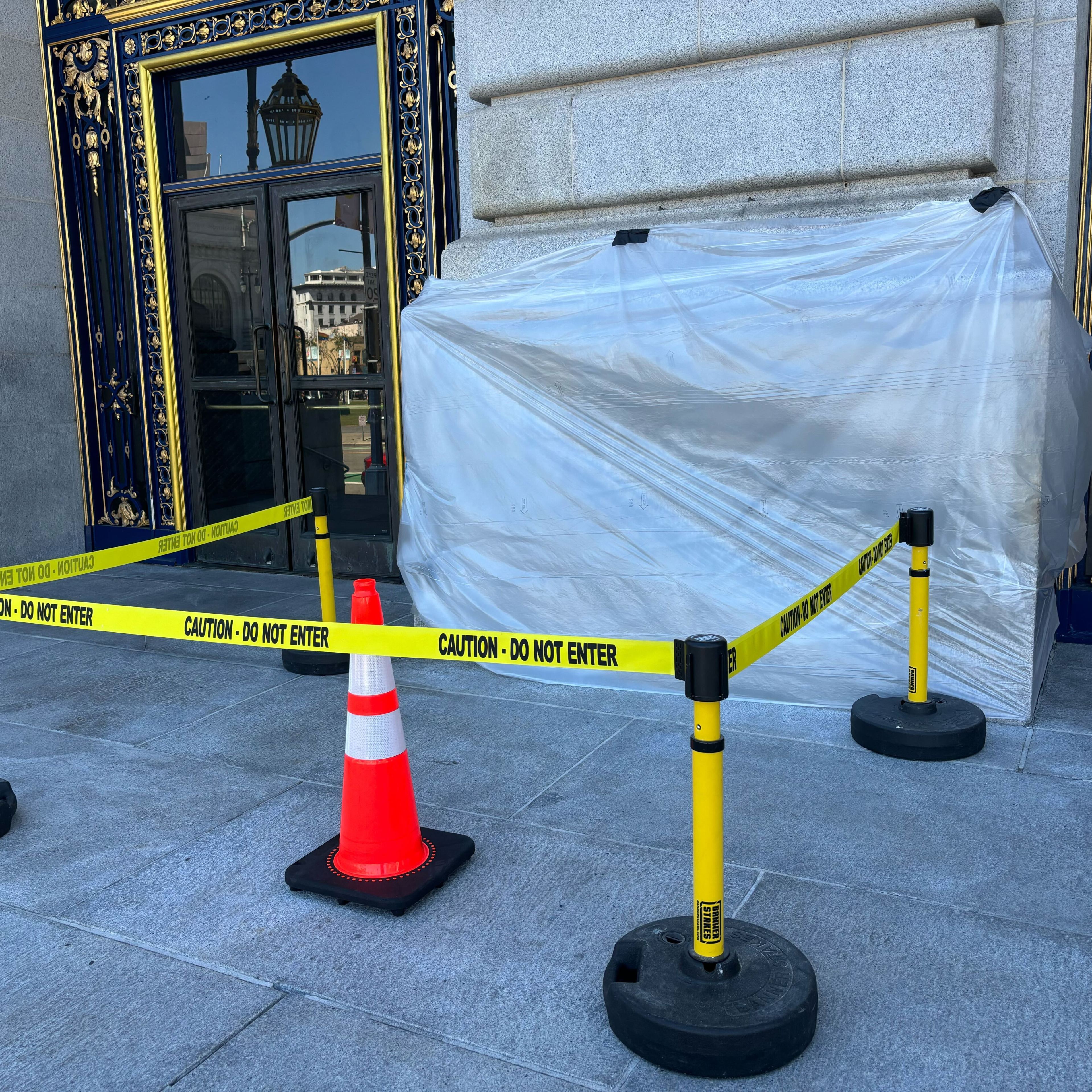 A doorway is blocked by "Caution - Do Not Enter" tape and a traffic cone. A large object covered in plastic is situated next to the door.