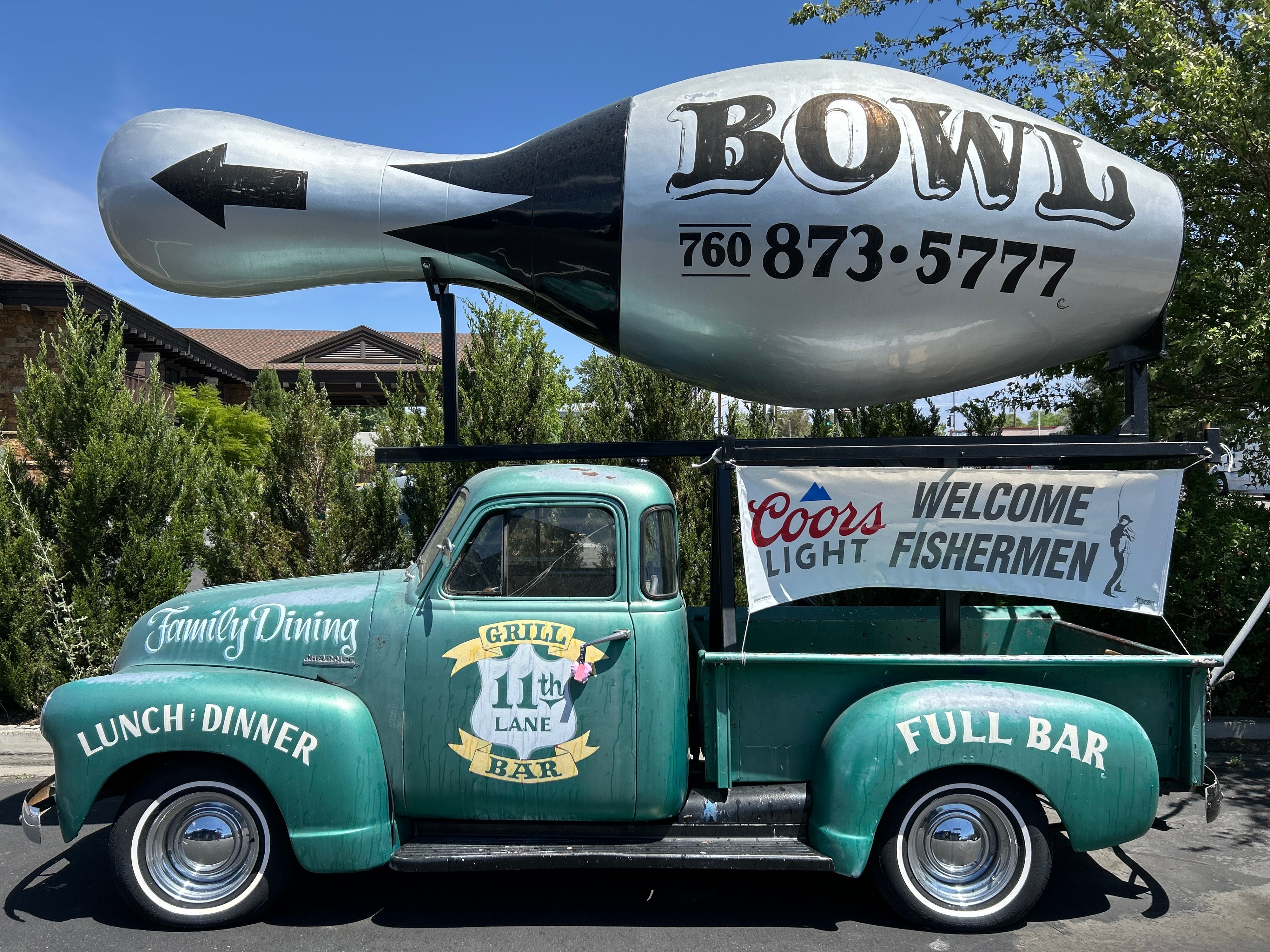 A vintage green truck is parked with a large bowling pin sign on top, advertising &quot;BOWL&quot; and a phone number. The truck features &quot;Family Dining,&quot; &quot;Grill 11th Lane Bar,&quot; and a banner saying &quot;Coors Light, Welcome Fishermen.&quot;