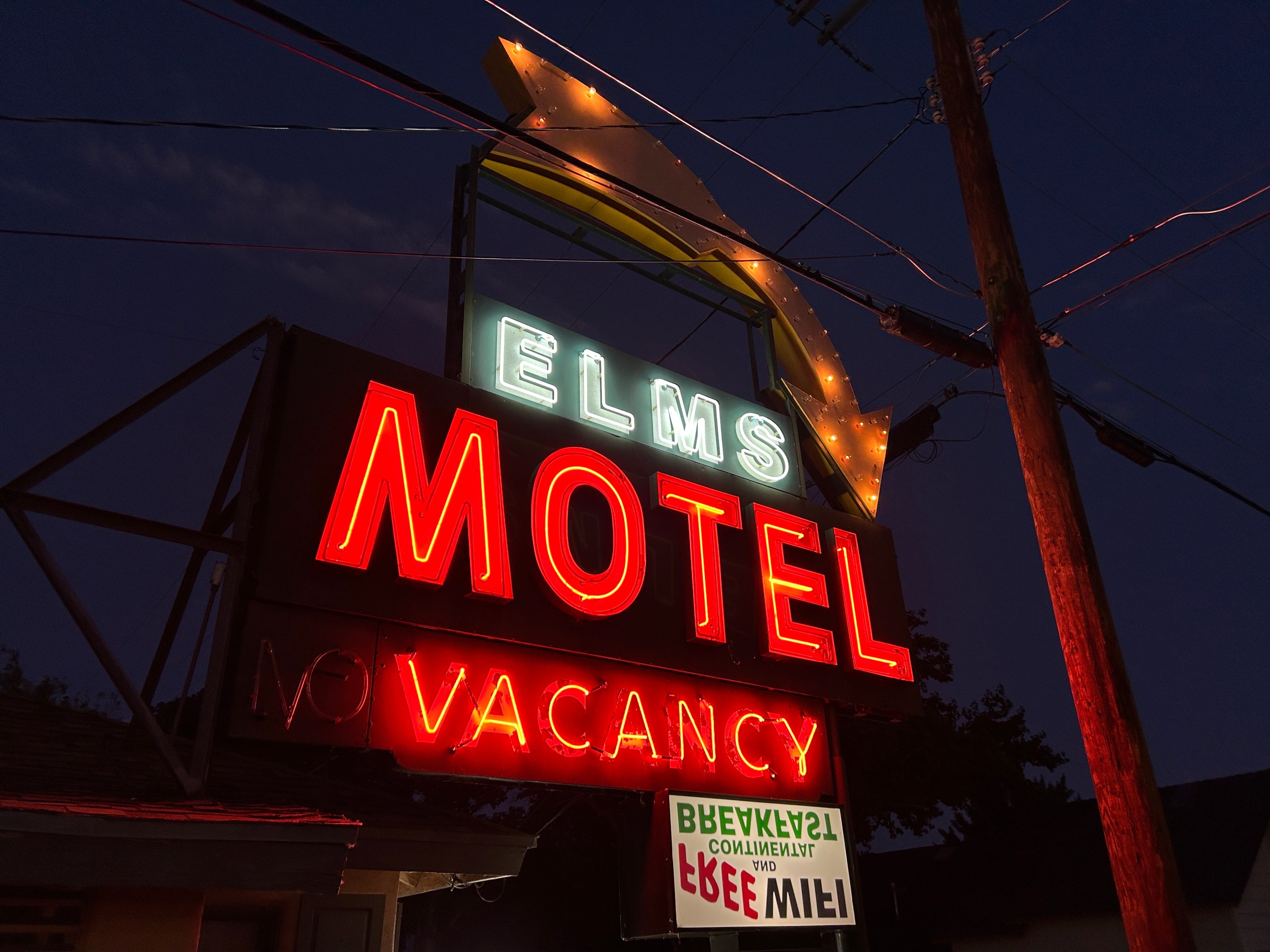 A brightly-lit neon sign reads &quot;Elms Motel.&quot; The word &quot;Vacancy&quot; is illuminated in red beneath the main sign, and a lit arrow points downwards.