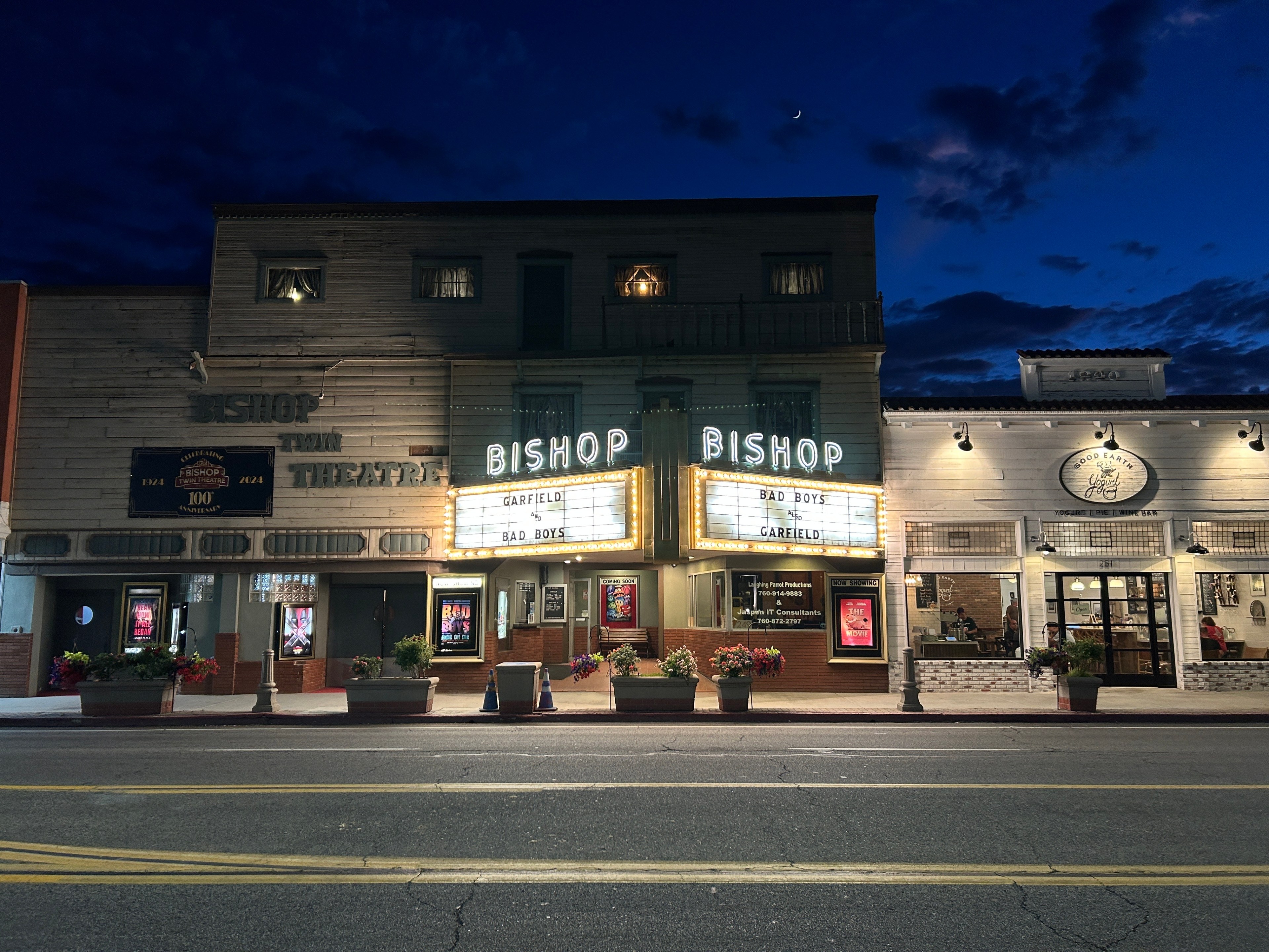 The image shows a lit-up historic theater named &quot;Bishop Twin Theatre&quot; at night. The marquee displays &quot;Garfield&quot; and &quot;Bad Boys.&quot; Next door is a cafe with several patrons inside.