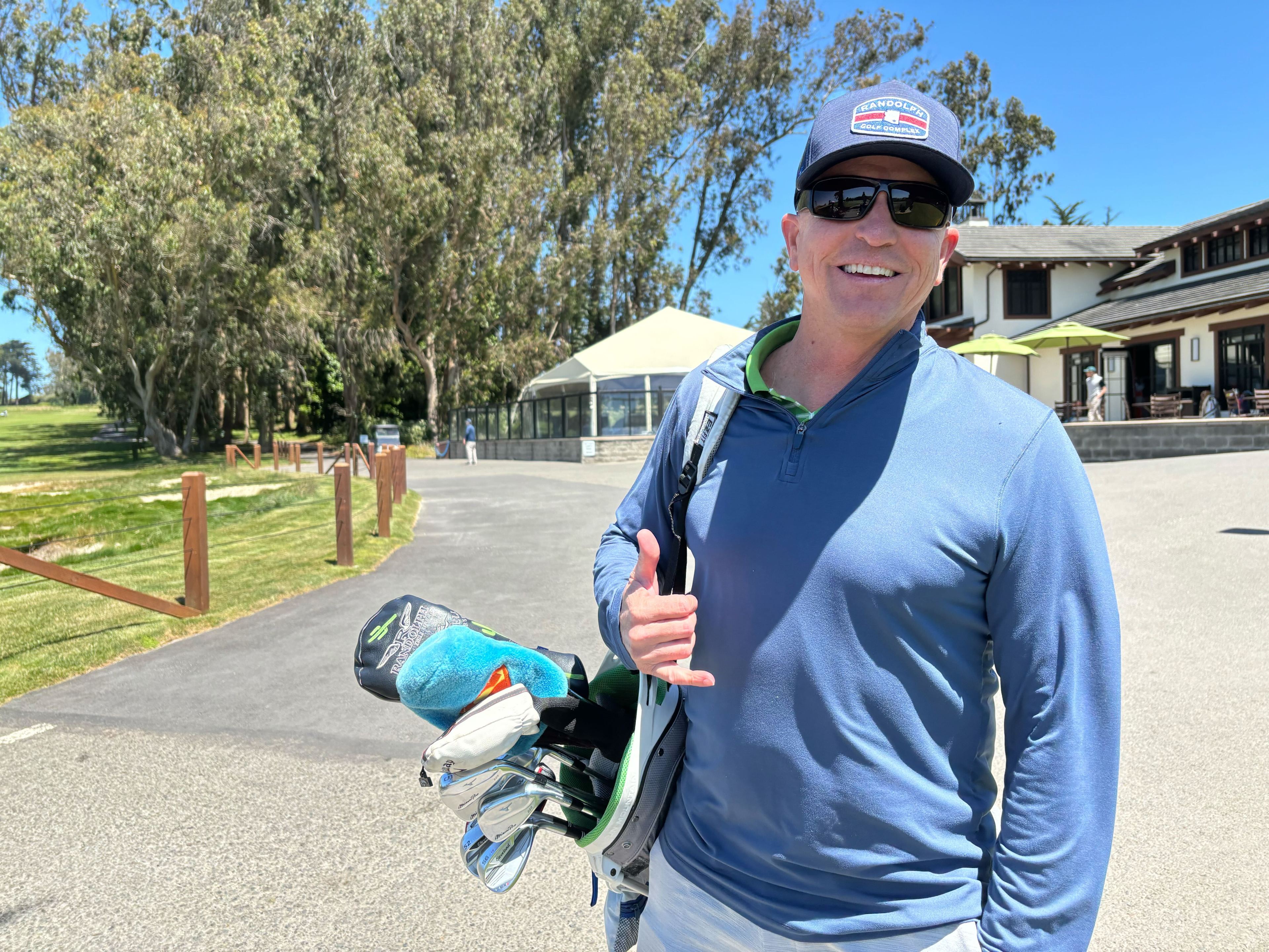 A golfer wearing a hat and sunglasses holds a bag of clubs over his shoulder.