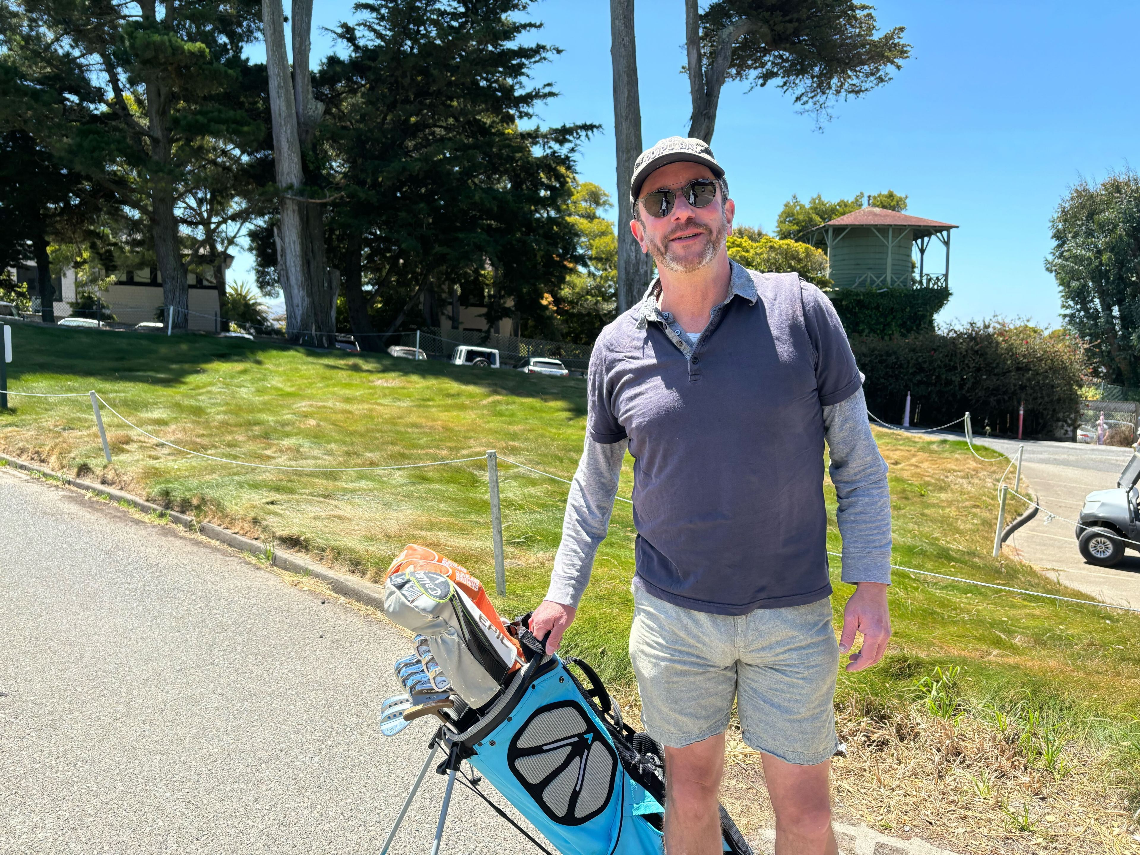 A golfer holds a bag of clubs on a sunny day at a golf course.