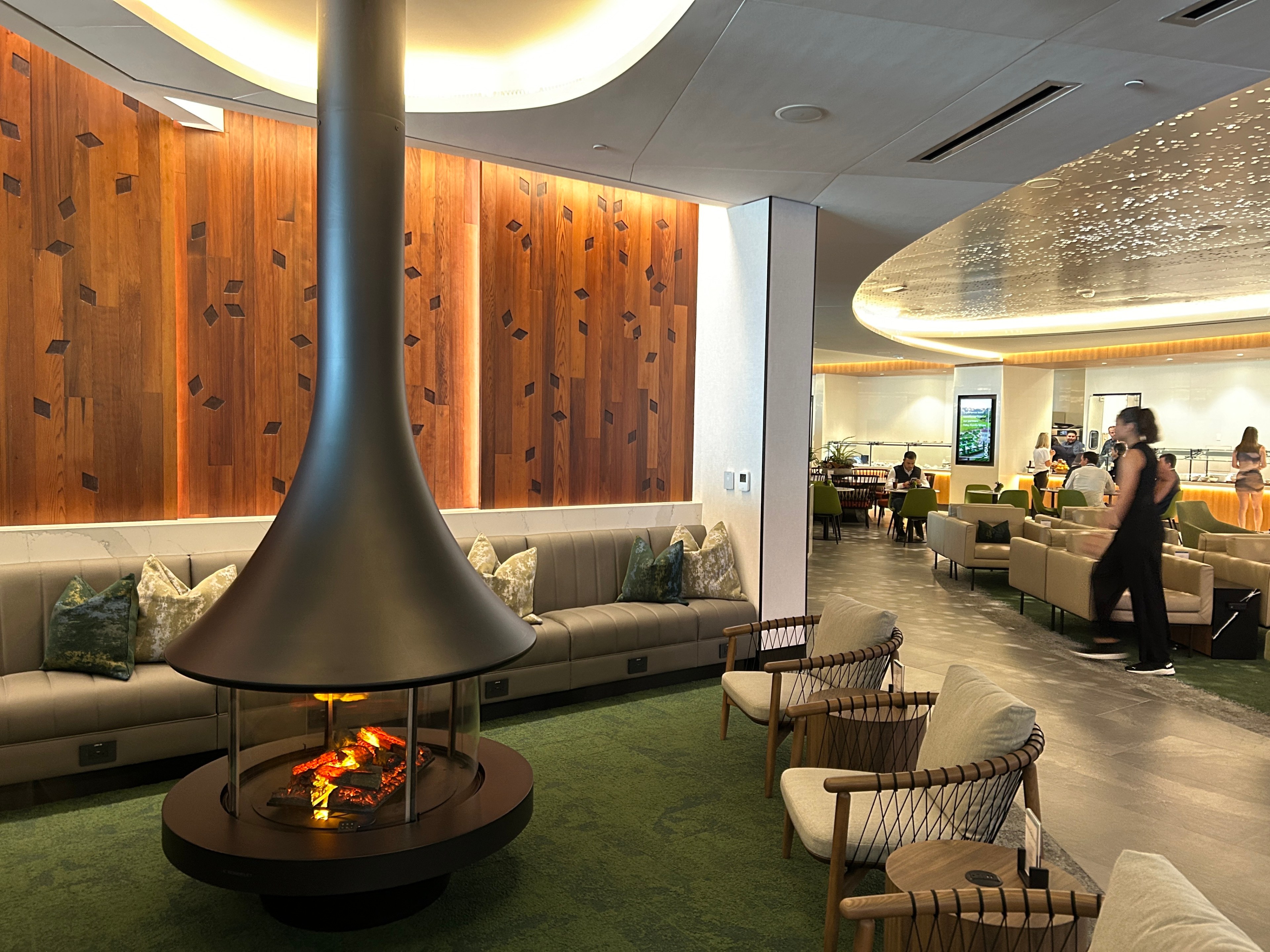 The image showcases a modern lounge area with a central cone-shaped fireplace, surrounded by seating options, including sofas and armchairs, with people dining in the background.
