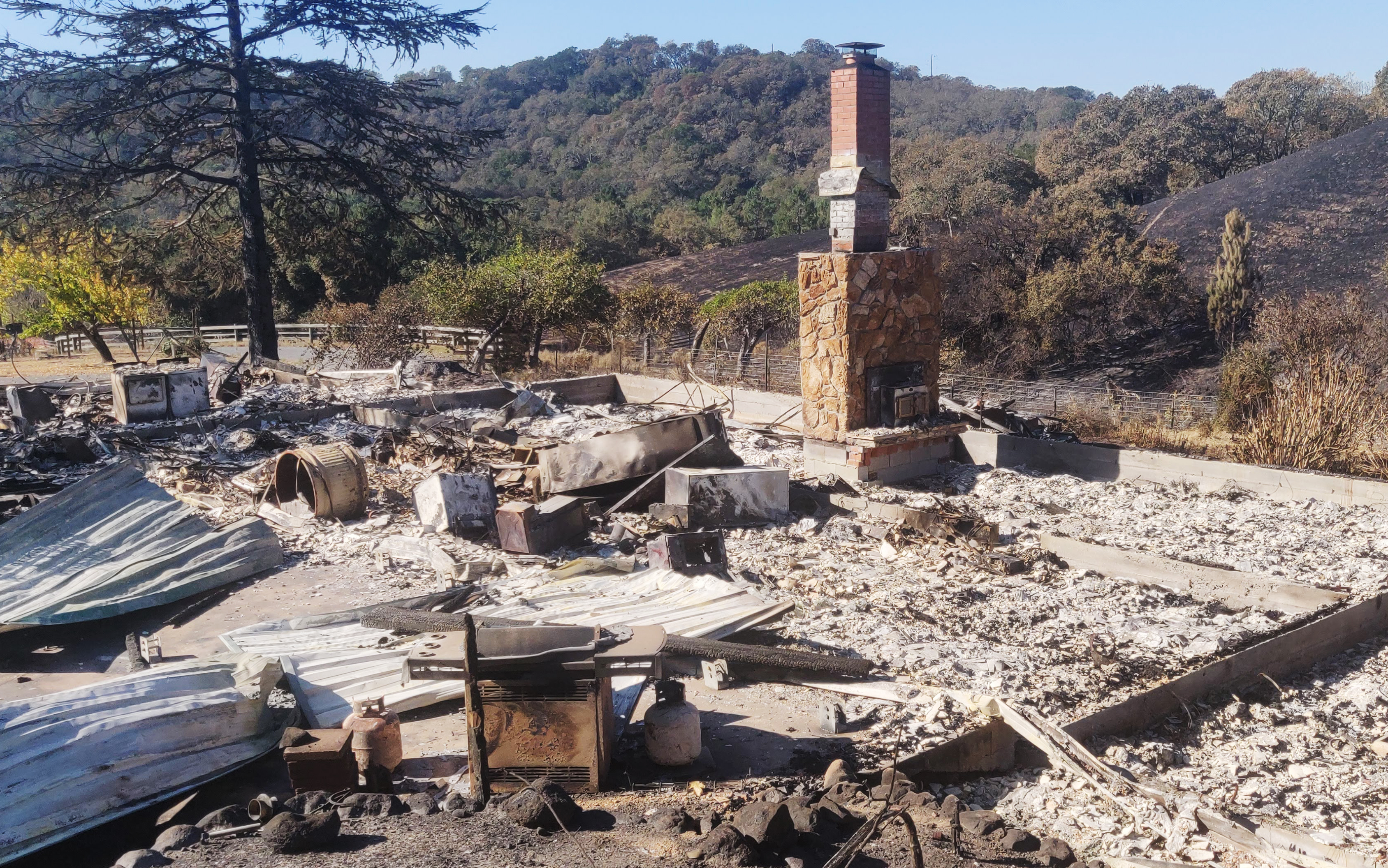 The image depicts the charred remains of a house after a fire, with rubble scattered on the ground and a brick chimney still standing amidst the destruction. A tree and hilly landscape provide the backdrop.