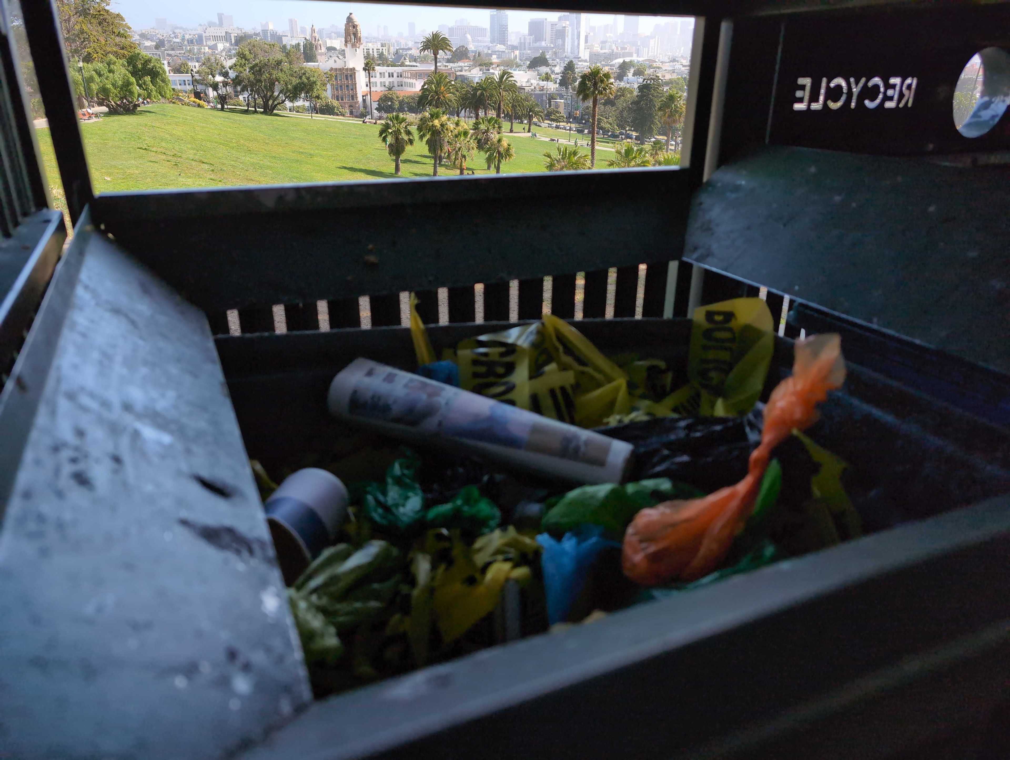The image shows a recycling bin filled with various items and a view of a park with palm trees, a tower, and a cityscape in the background through the bin's opening.