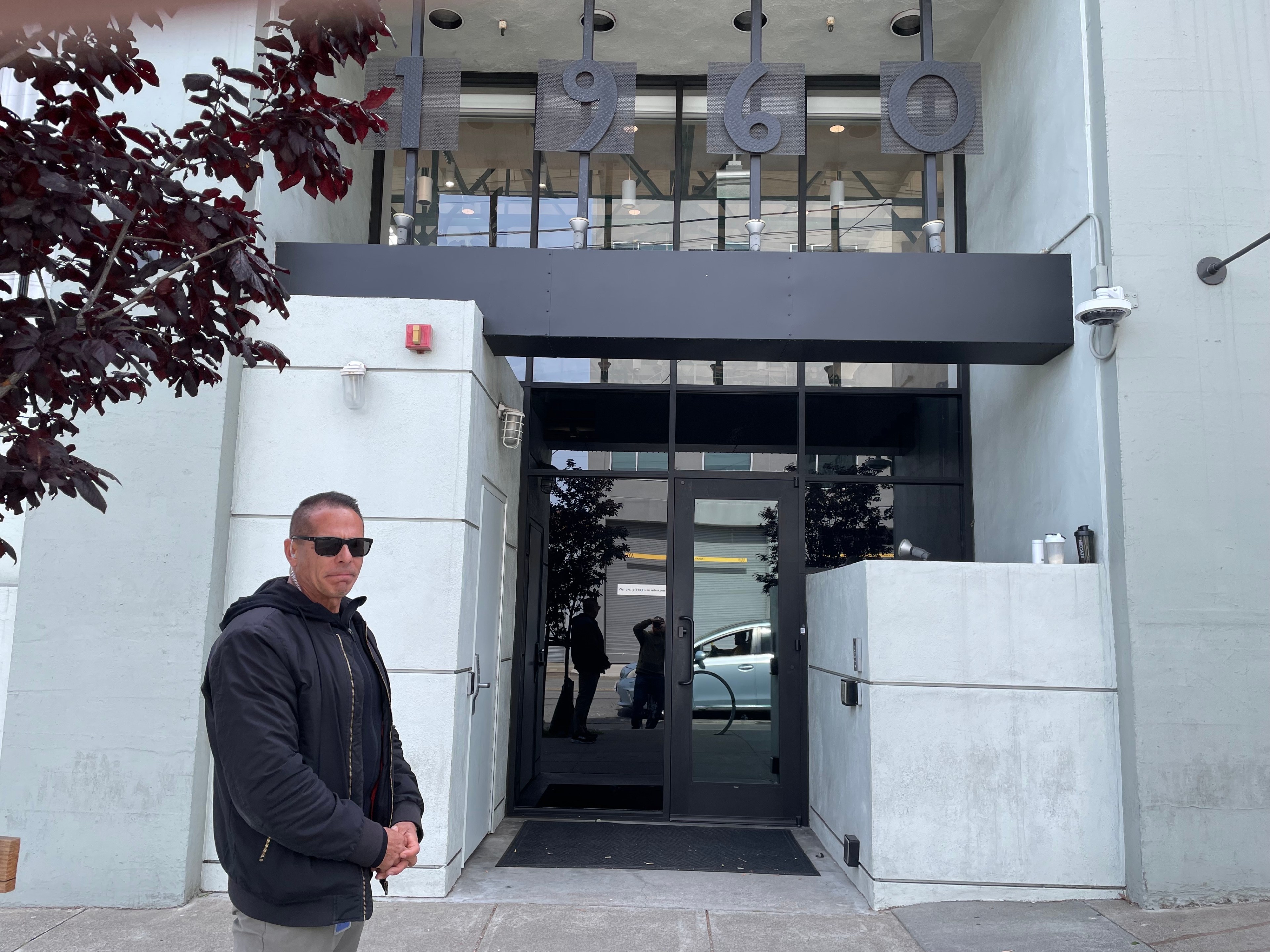 A man wearing sunglasses and an earpiece stands outside an office building.