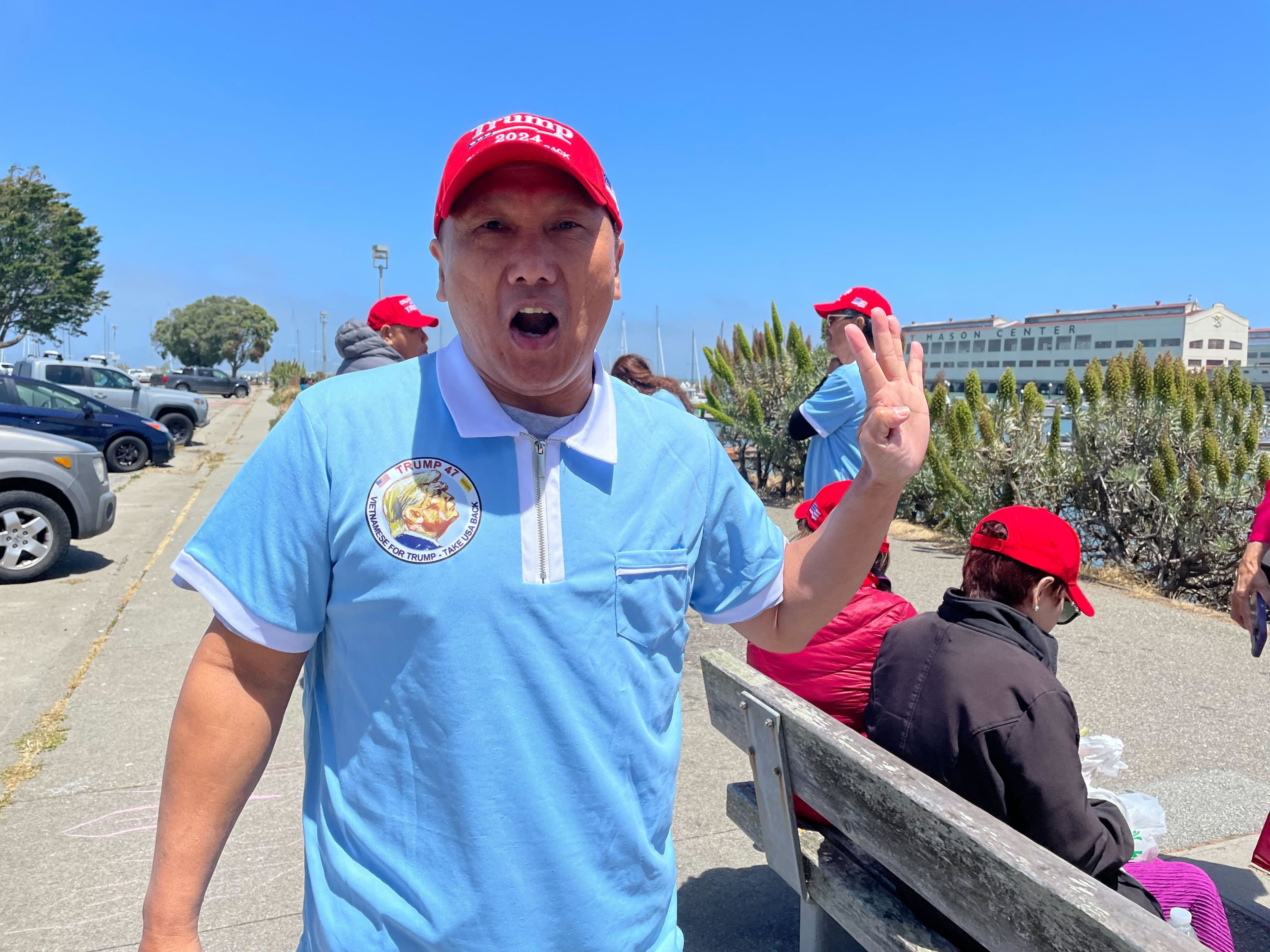 A man wearing a red &quot;Trump 2024&quot; hat and a blue shirt with a Trump badge gestures a &quot;peace&quot; sign near benches and a group of people also in red hats.