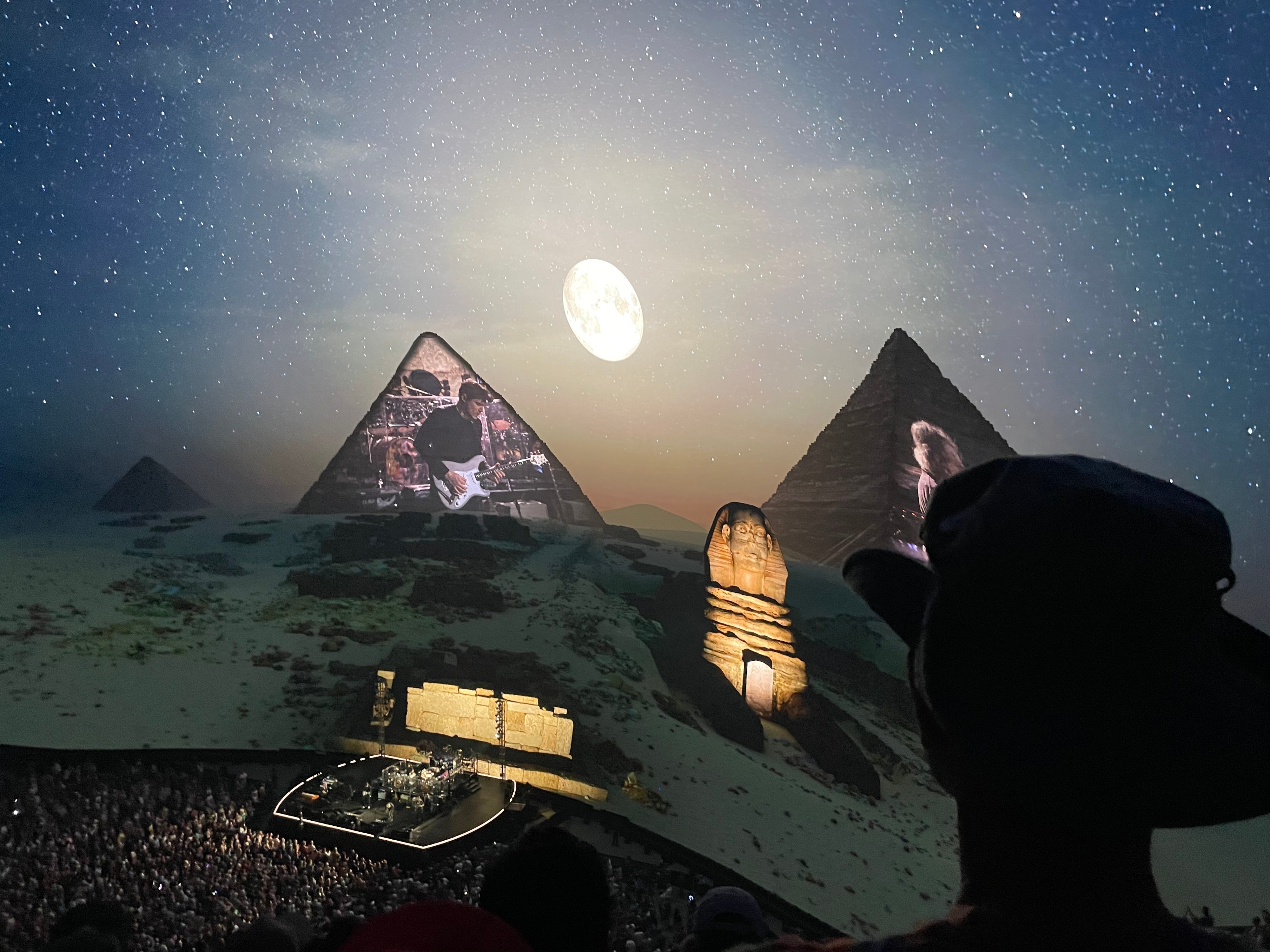A night concert features a stage below the Pyramids and Sphinx, with projected images, a full moon, and a starry sky, viewed by an audience in the foreground.