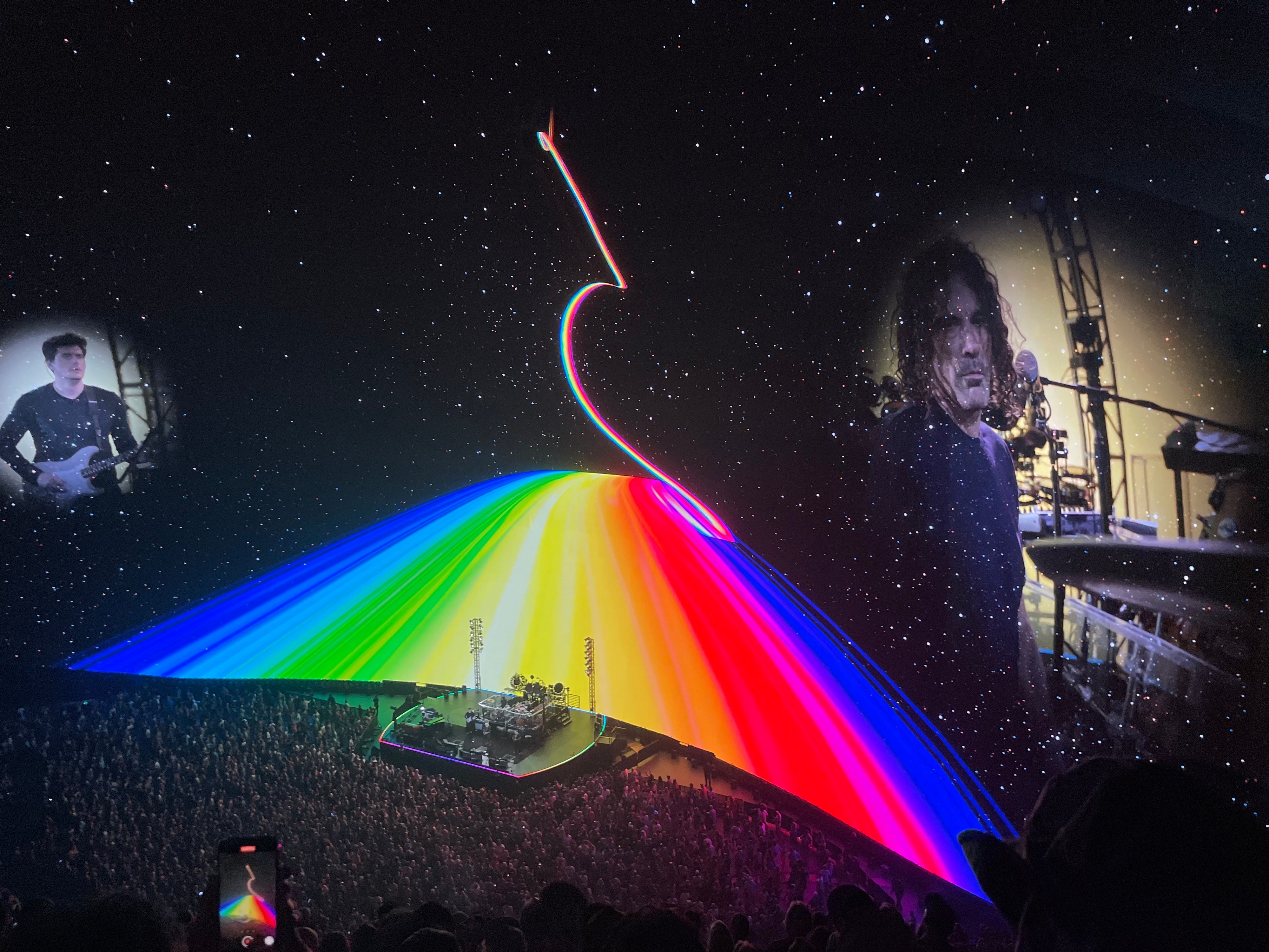 A concert stage with a large, colorful, rainbow-like light display and outer space background features images of two musicians projected on either side.