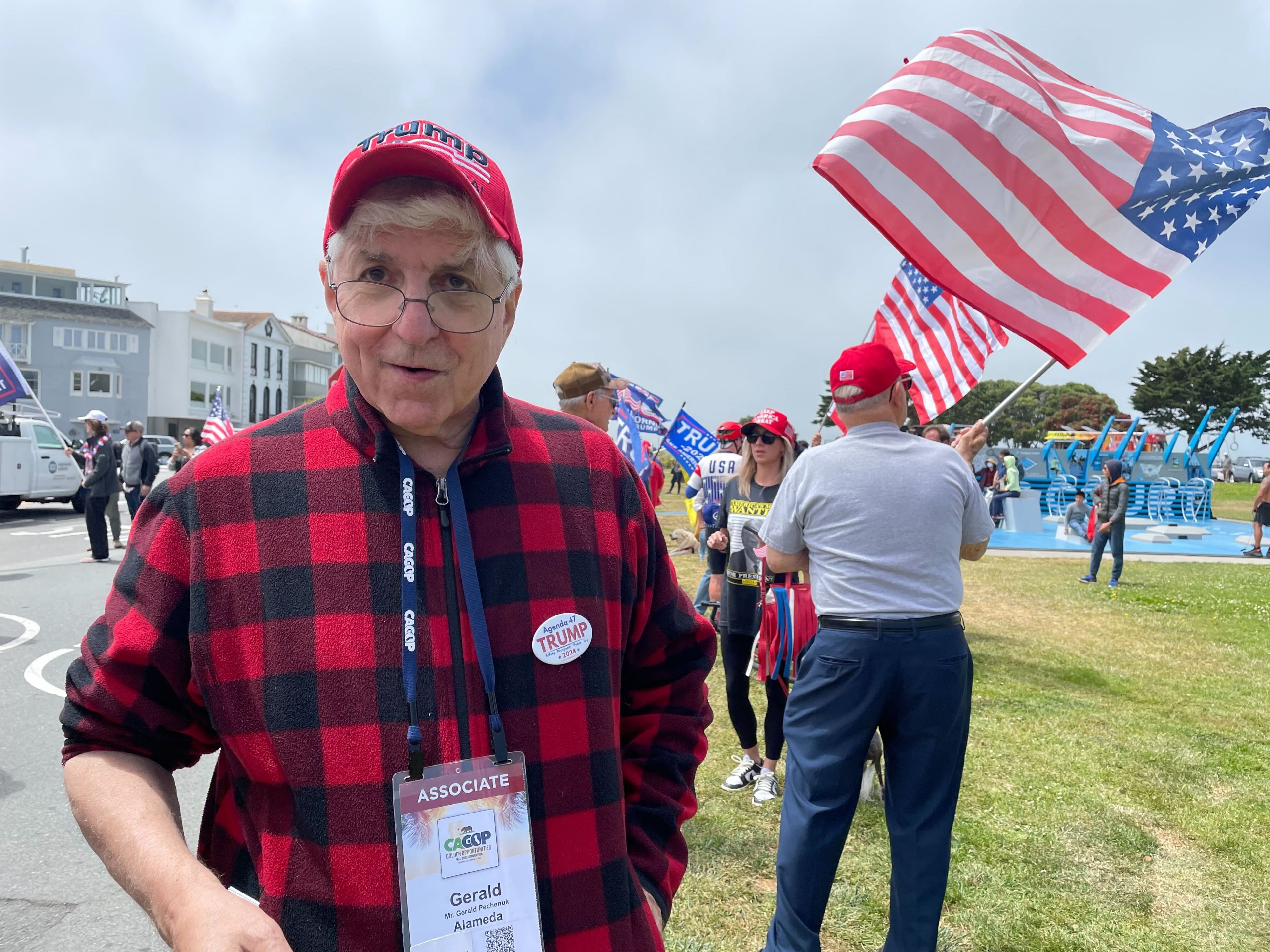 An older man in a red plaid shirt and red cap stands in the foreground, with numerous American flags and people in the background at an outdoor event.
