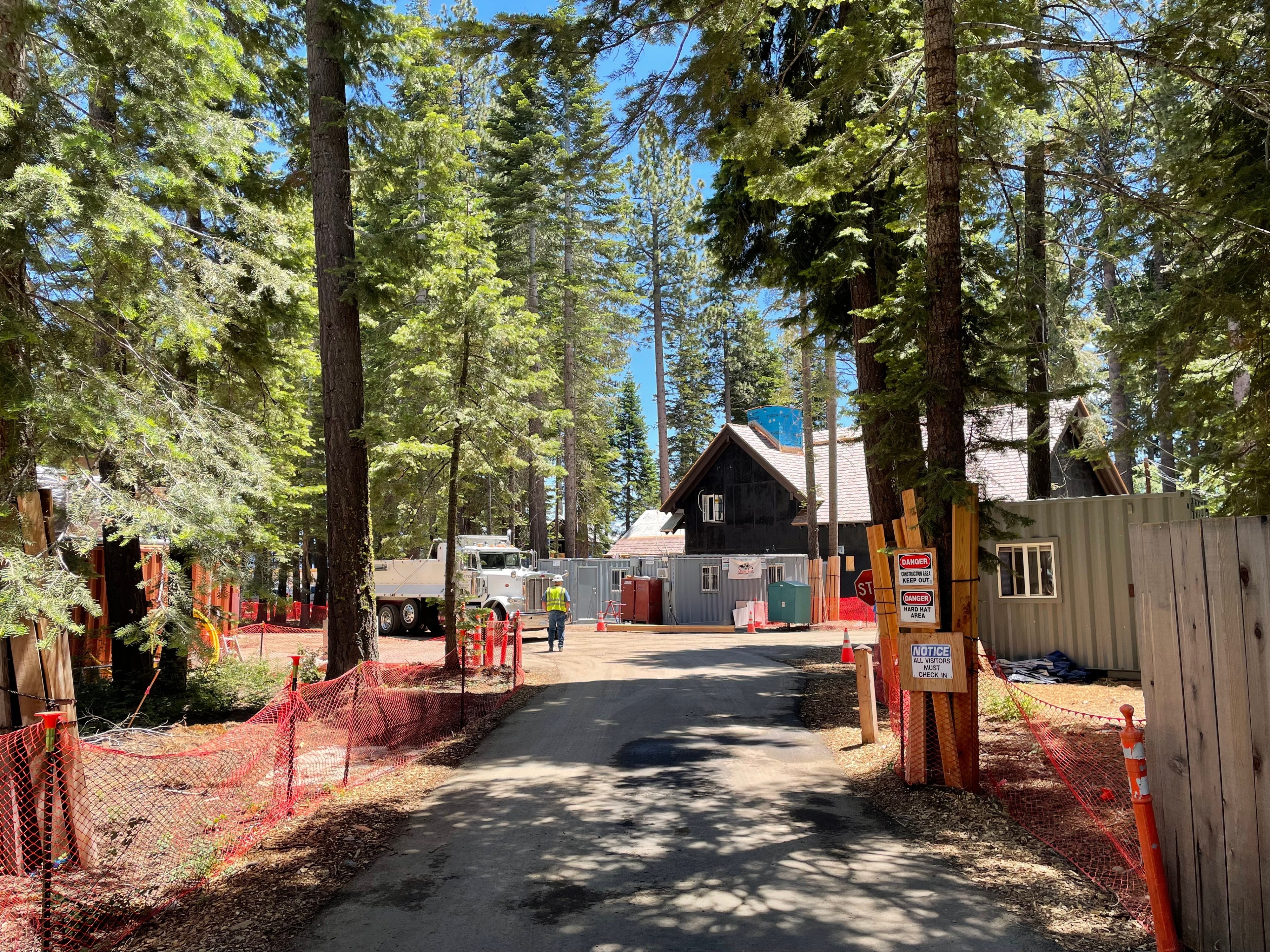 A wooded construction site features scattered homes, a parked truck, red safety fencing, various signs, and a clear sunny sky.