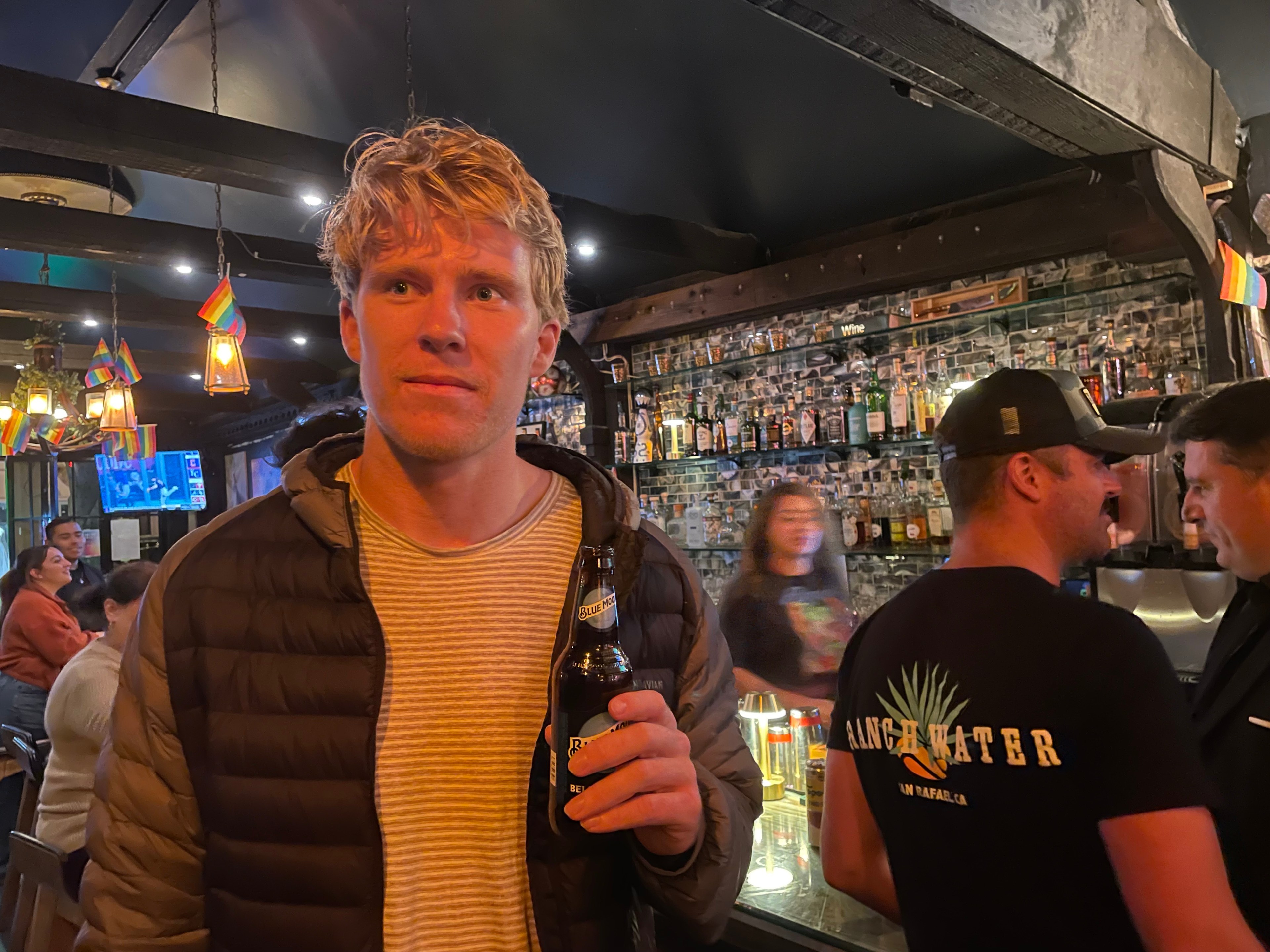 A man with blond hair, wearing a striped shirt and puffy jacket, holds a bottle in a bar. The background shows people, shelves of liquor, and pride flags.