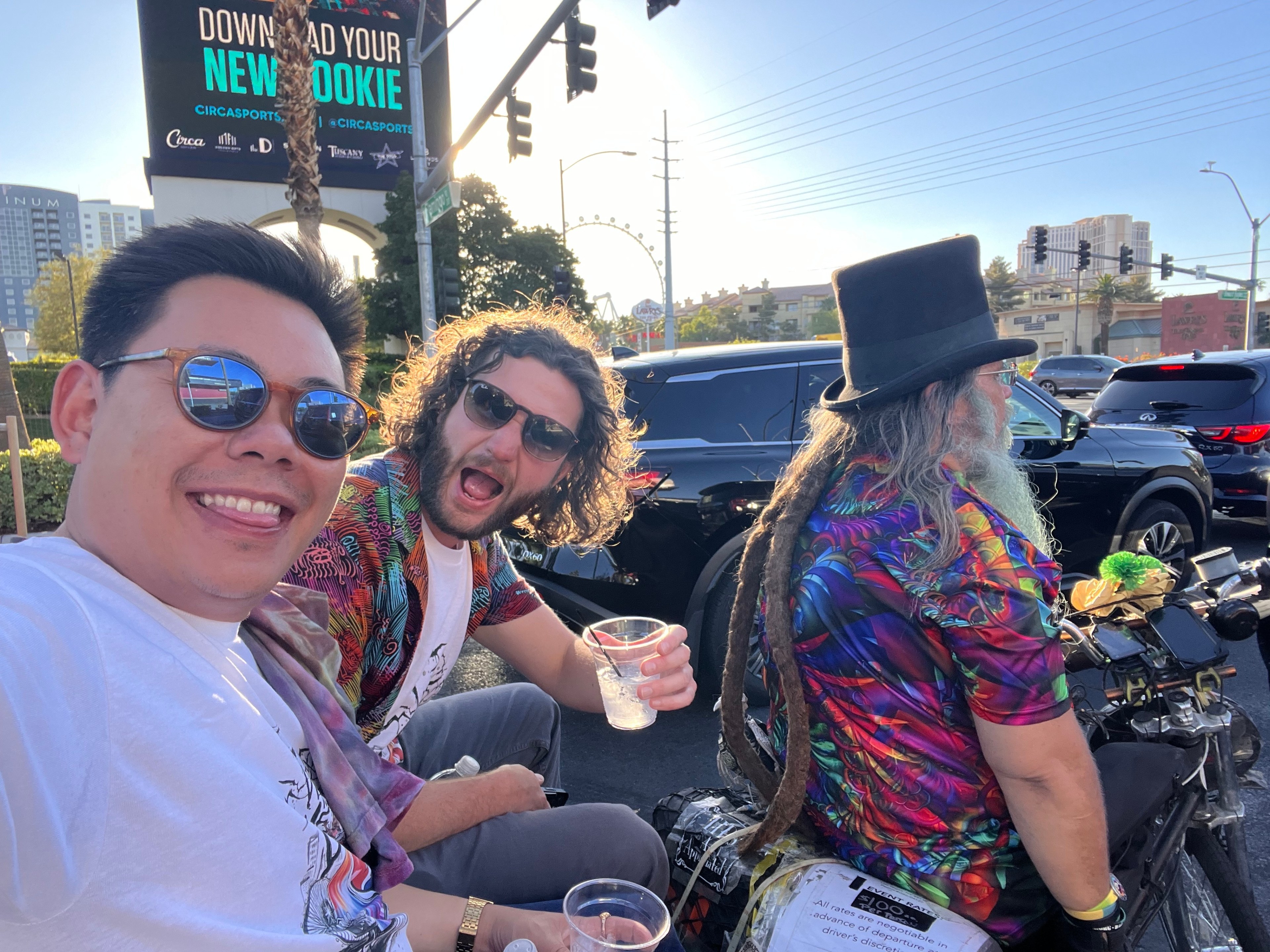 Three men in vibrant attire are enjoying drinks on a street corner; one has long dreadlocks and a top hat. Behind them are cars and a large billboard.