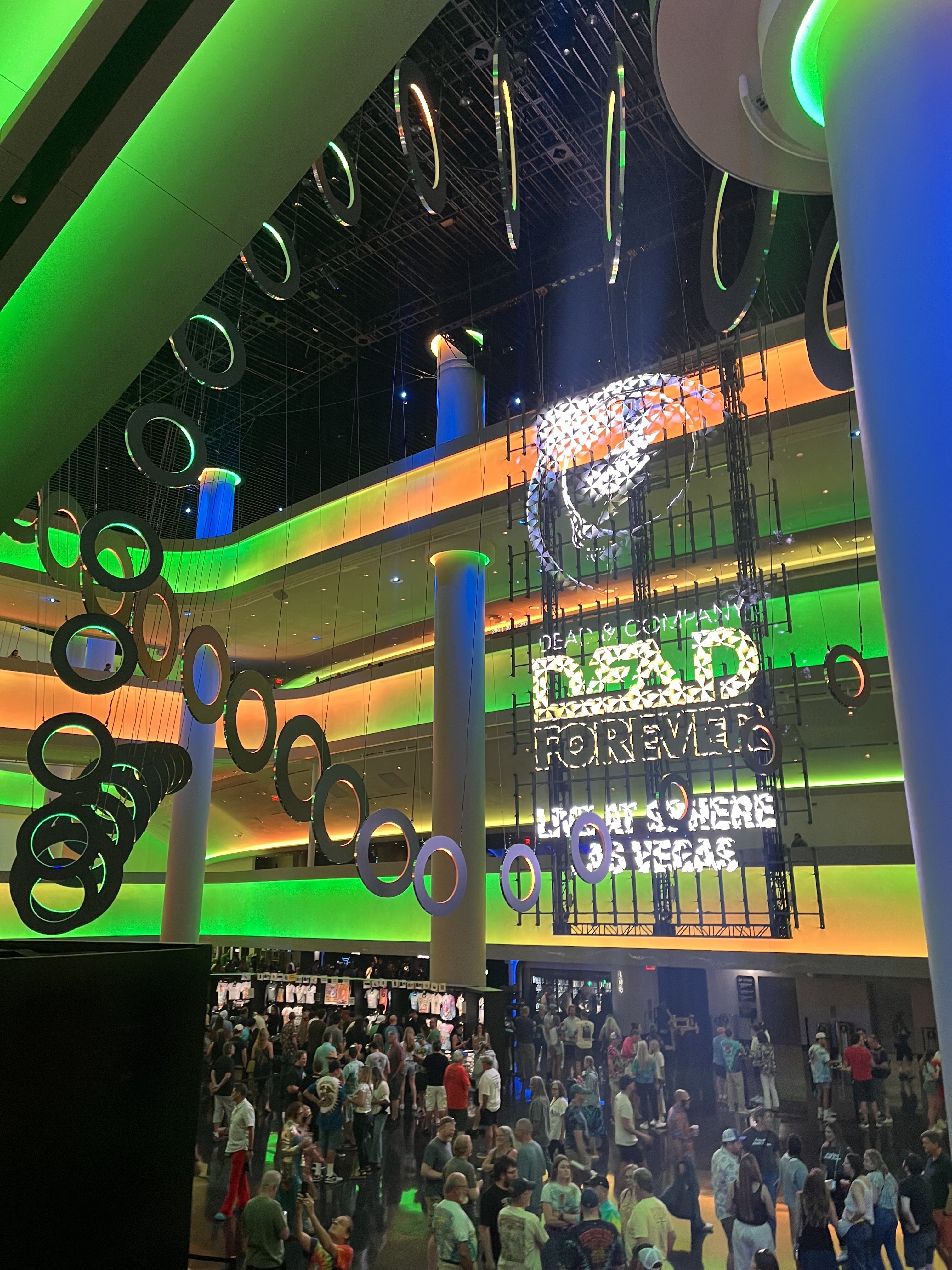 The image shows a large indoor event space with colorful lighting, circular hanging decorations, a crowd of people below, and a large illuminated sign that reads “Dead &amp; Company, The Final Tour.”