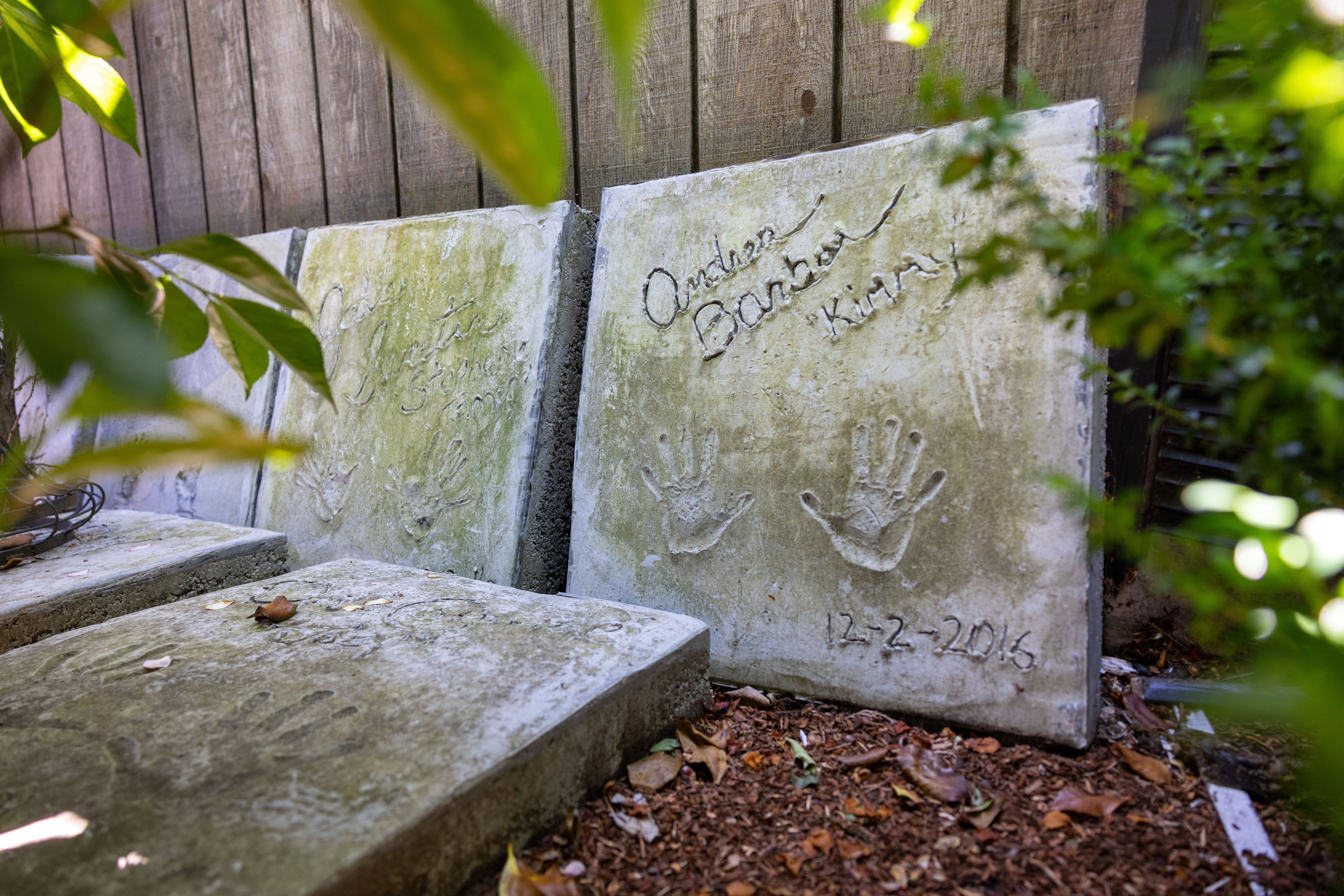 Concrete slabs with handprints and names etched in them rest against a wooden fence, surrounded by foliage. One slab is dated 12-2-2016.