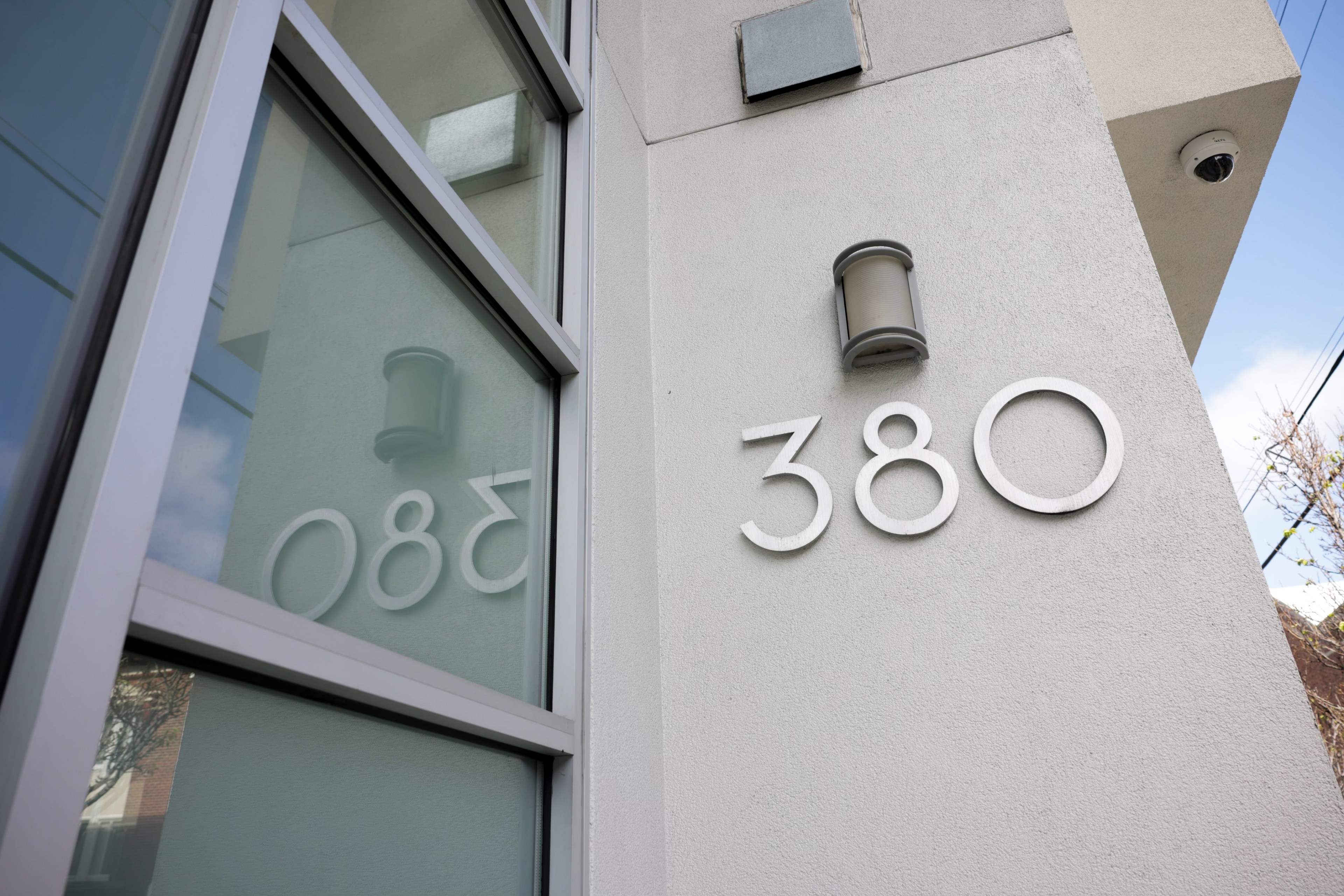 The image shows a modern building corner with the number &quot;380&quot; on the wall. A light fixture is mounted above the number, and a reflection is seen in a nearby window.