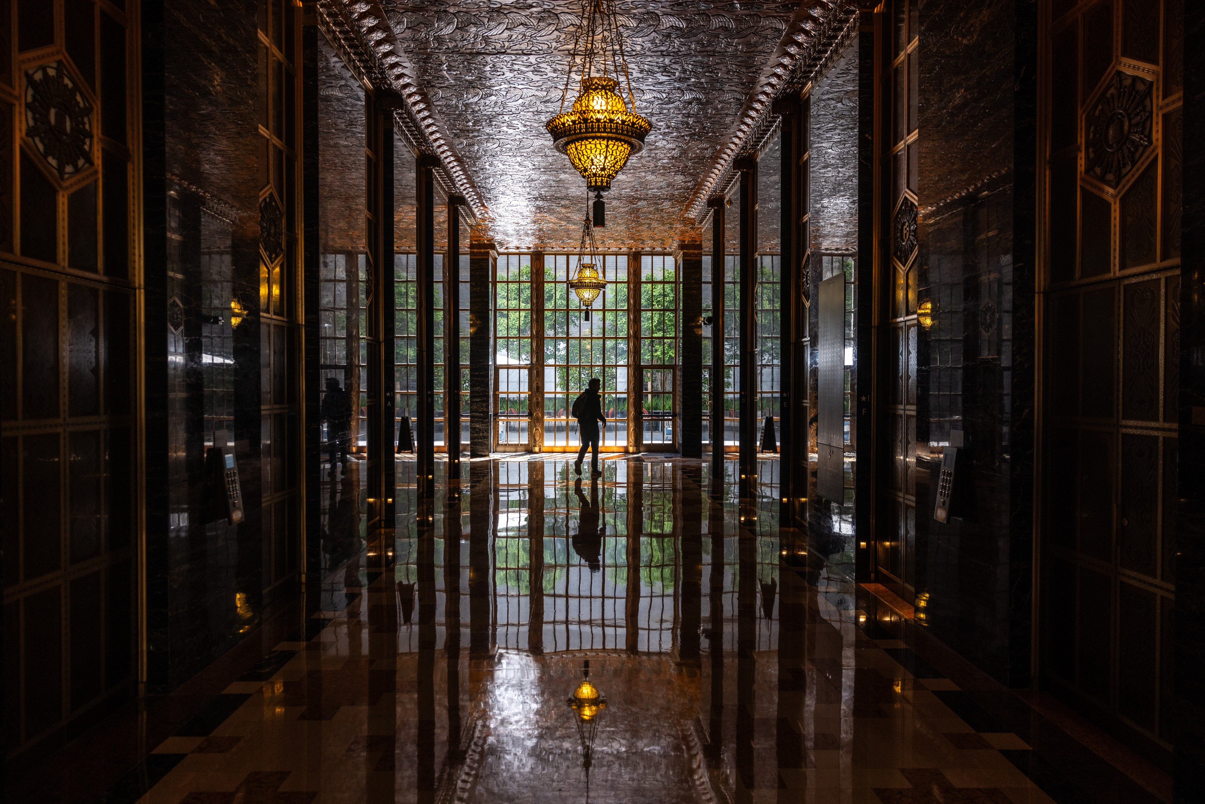 A grand hallway features intricate chandeliers, glossy reflective floors, and large windows at the end, framing a lone figure walking toward the light.