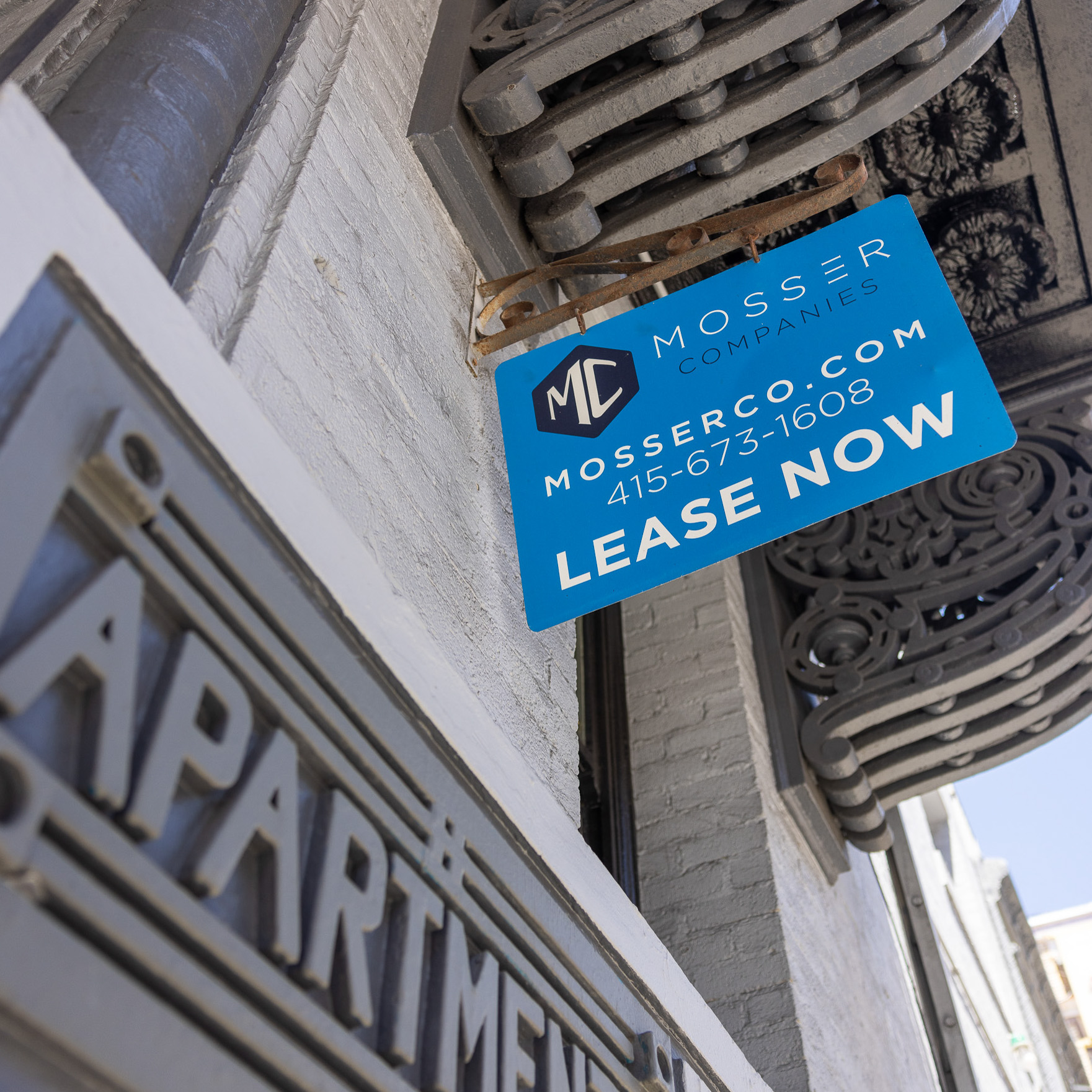 A blue sign attached to a building reads &quot;Mosser Companies&quot; with their web address, phone number, and &quot;Lease Now.&quot; The perspective shows ornate structural details and part of a sign saying &quot;Apartment.&quot;