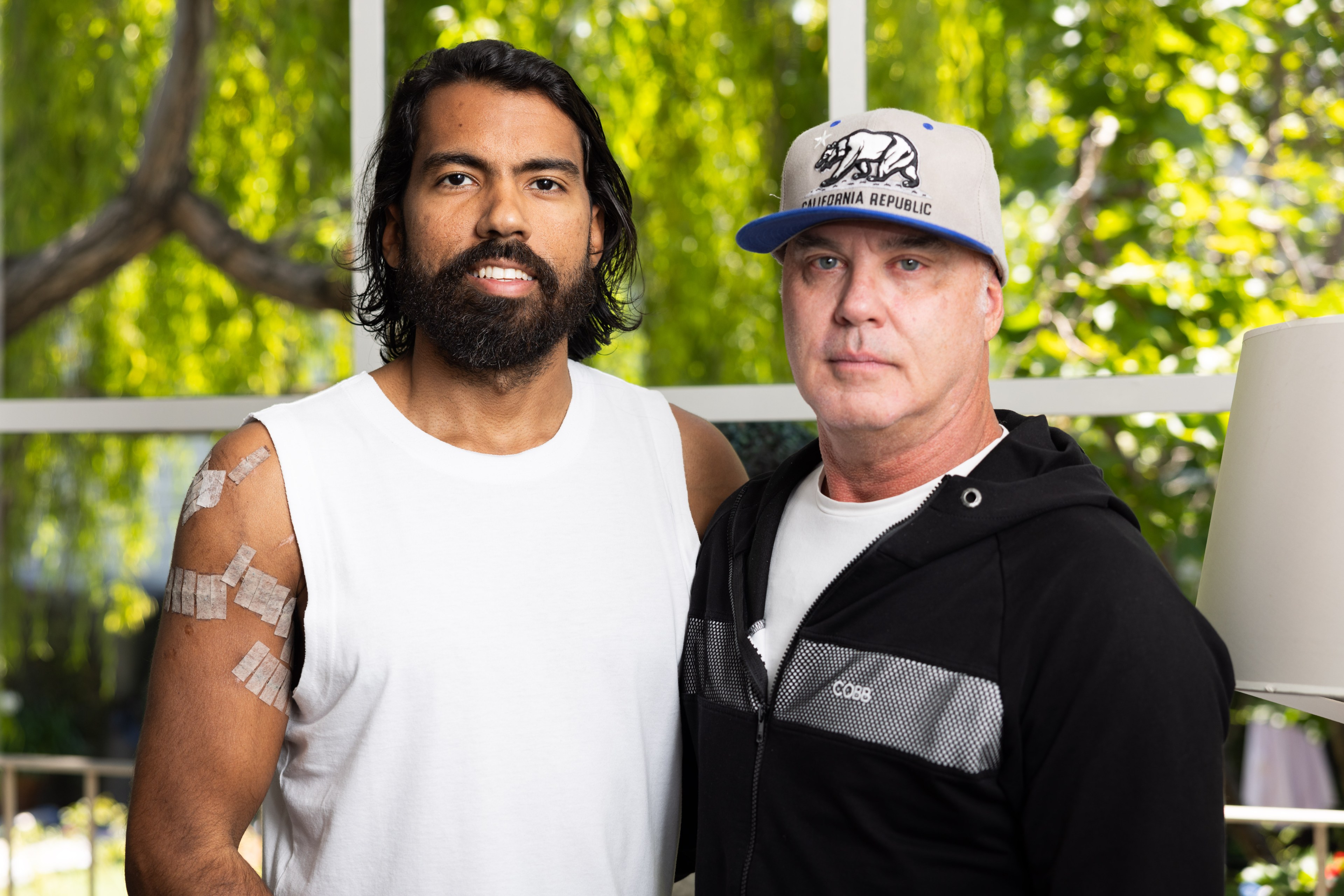 Two men standing together; the left man has long hair and a beard, with medical tape on his arm, wearing a white sleeveless shirt. The right man wears a cap and a black hoodie.