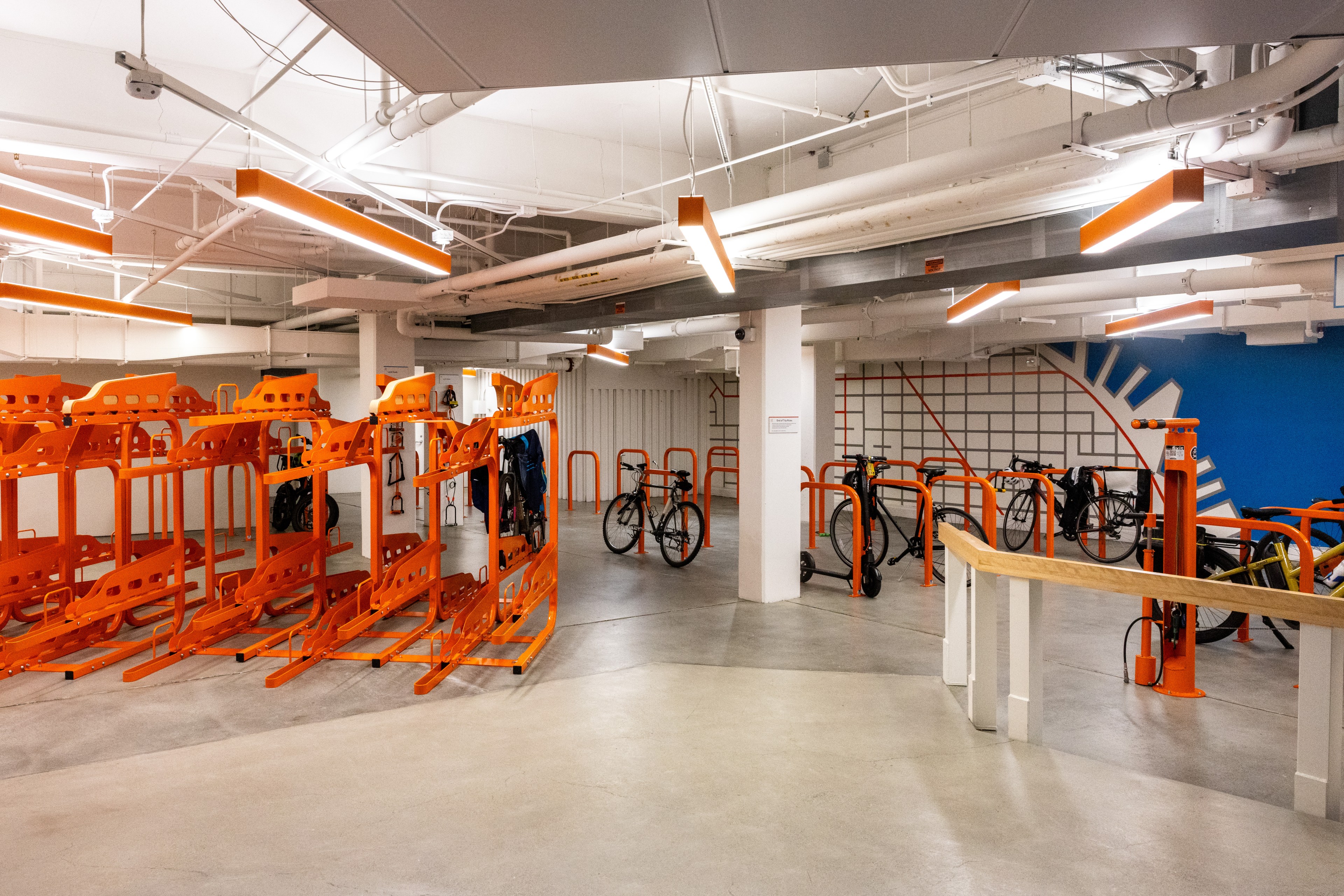 The image shows an indoor bicycle storage area with bright orange bike racks and hooks. Several bicycles are parked, and the space has a clean, modern design.