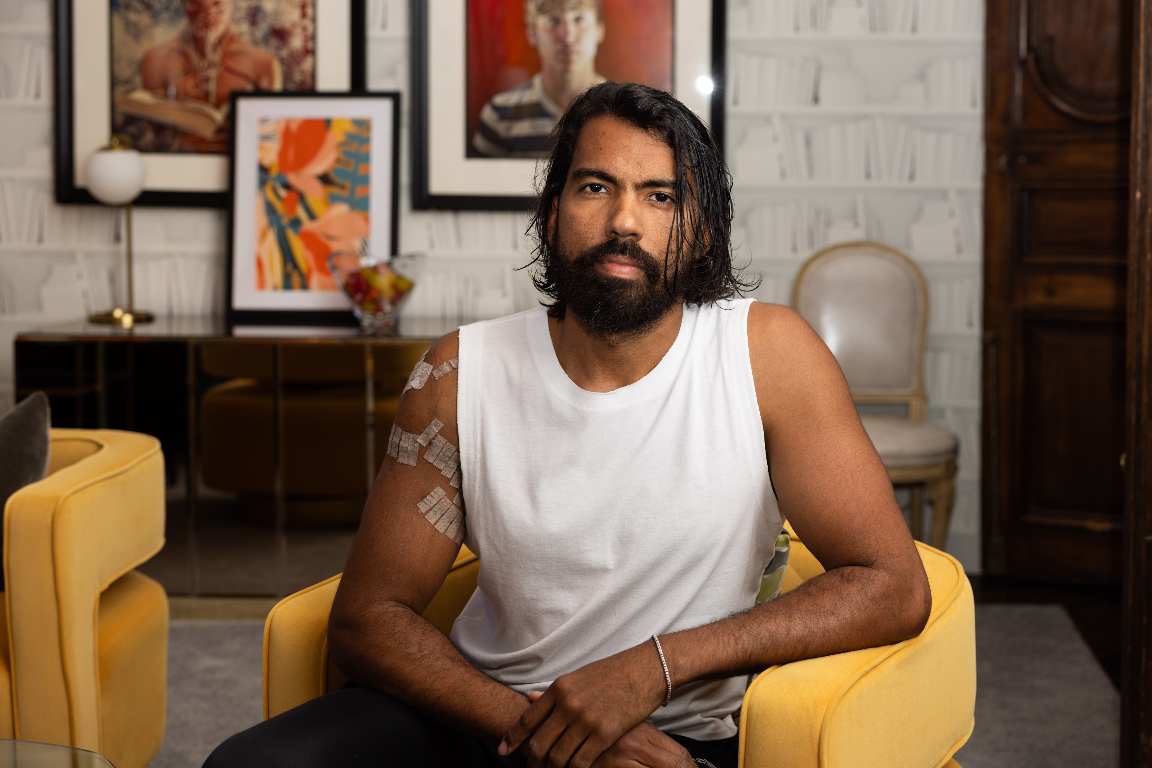 A bearded man with shoulder-length hair sits in a yellow chair, wearing a sleeveless white shirt with medical tape on his arm, in a room with framed art and cozy furnishings.