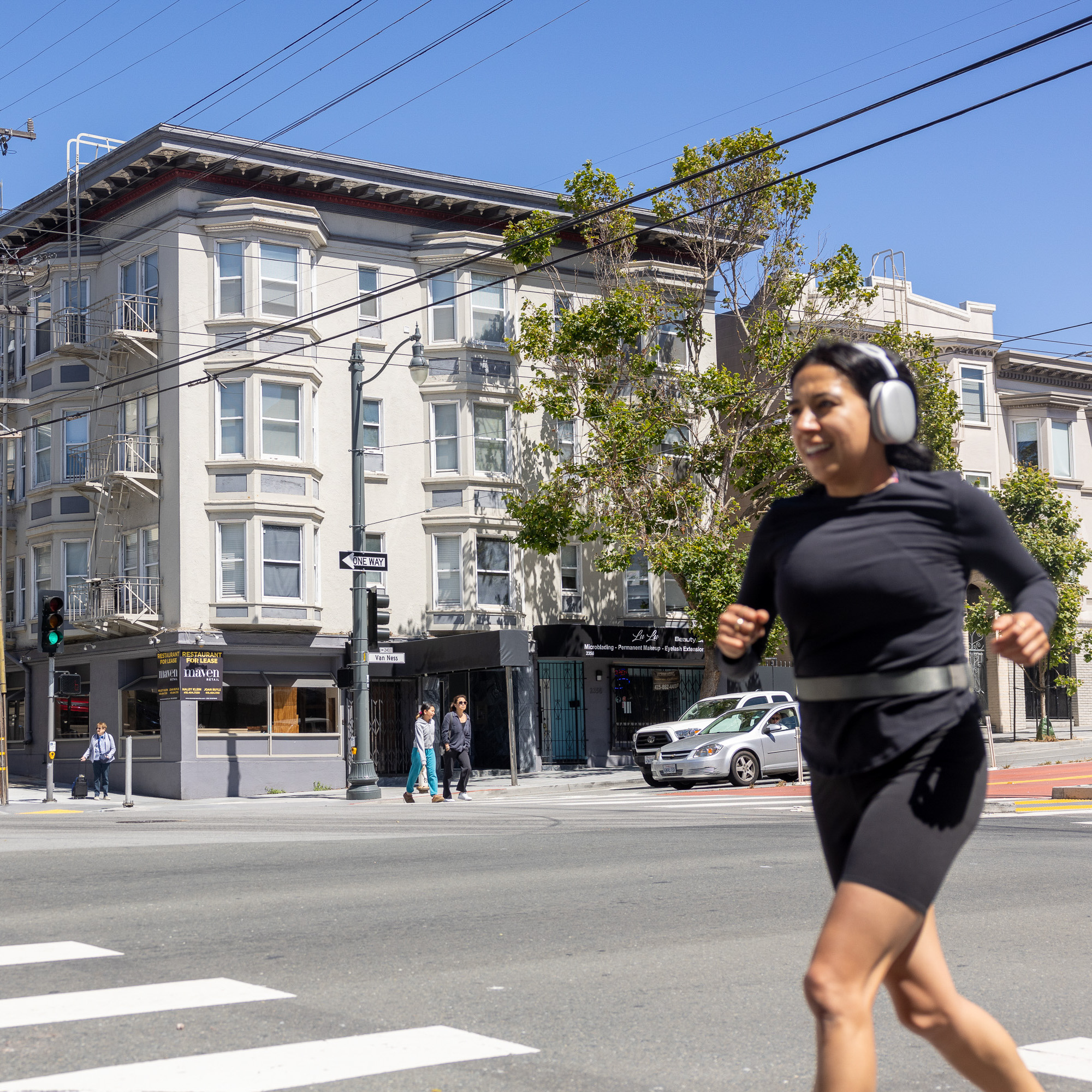 A woman with headphones jogs past a crosswalk on a sunny day, with a beige multi-story building, trees, and a few pedestrians in the background.