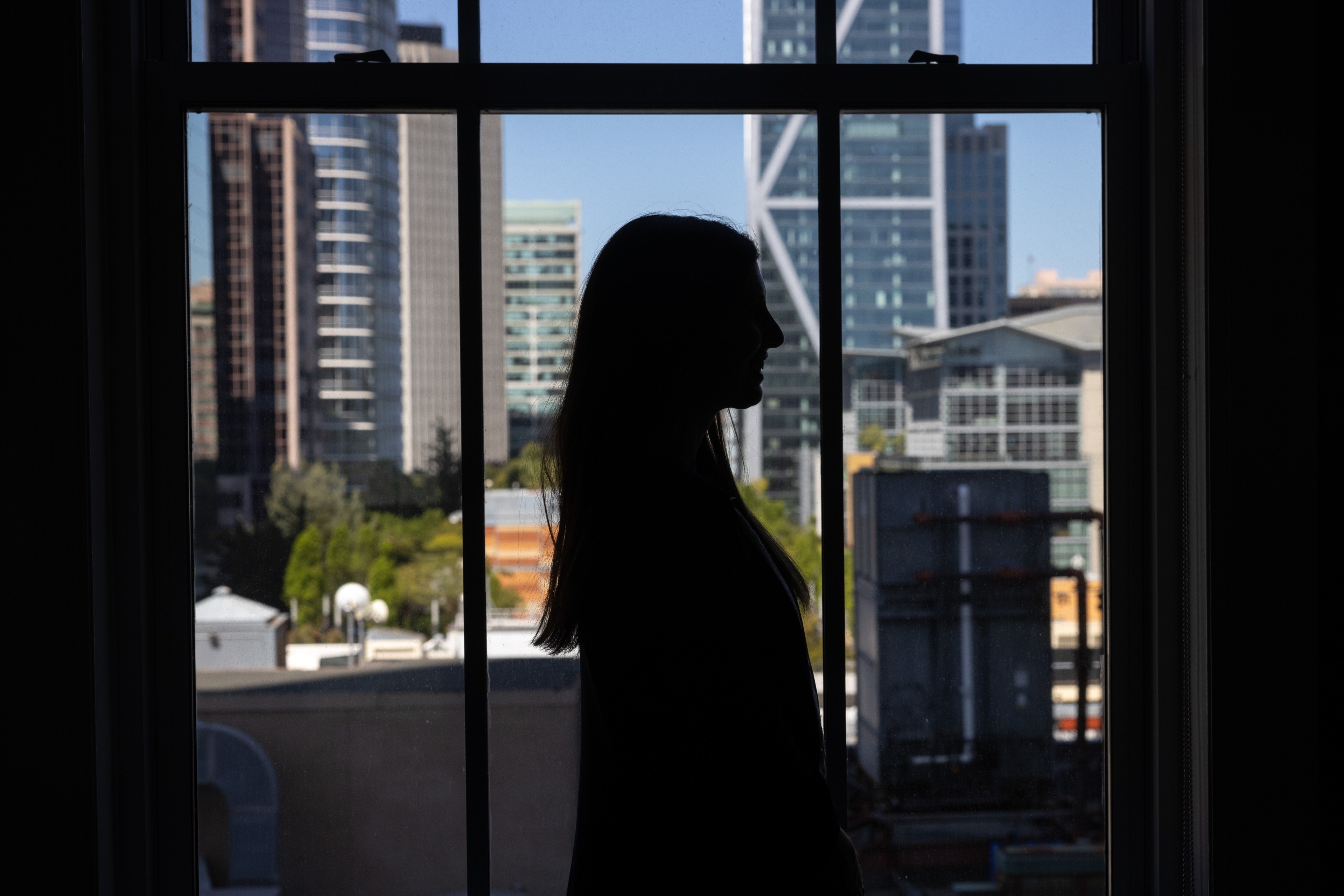 A silhouetted person stands in front of a large window with a city skyline comprising various modern high-rise buildings in the background.