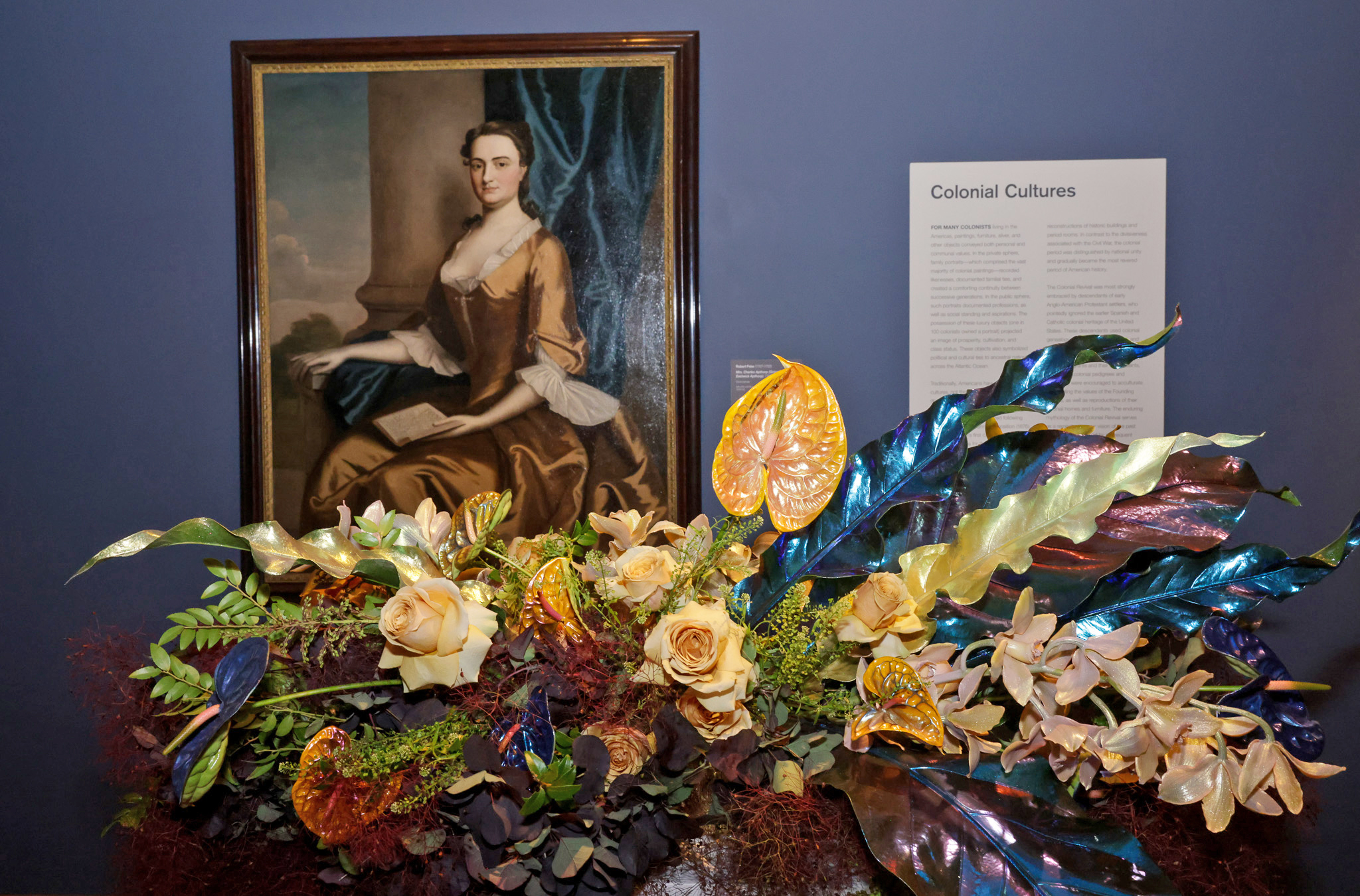 The image shows a framed portrait of a woman in historical attire, displayed on a blue wall with a floral arrangement of roses and glossy leaves beneath it.