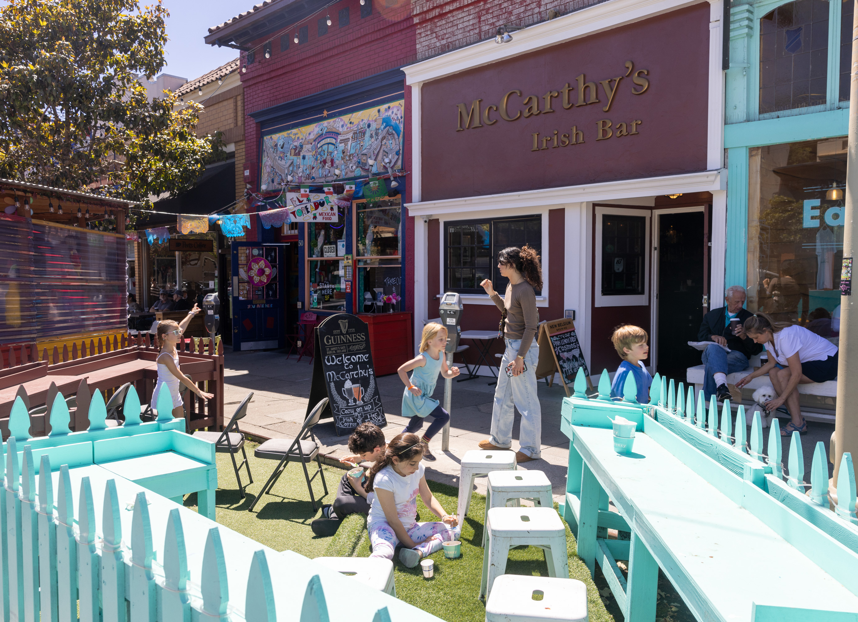 Children and adults socialize outside McCarthy's Irish Bar. Bright decor, a blue fence with benches, and colorful storefronts add vibrant energy to the street scene.