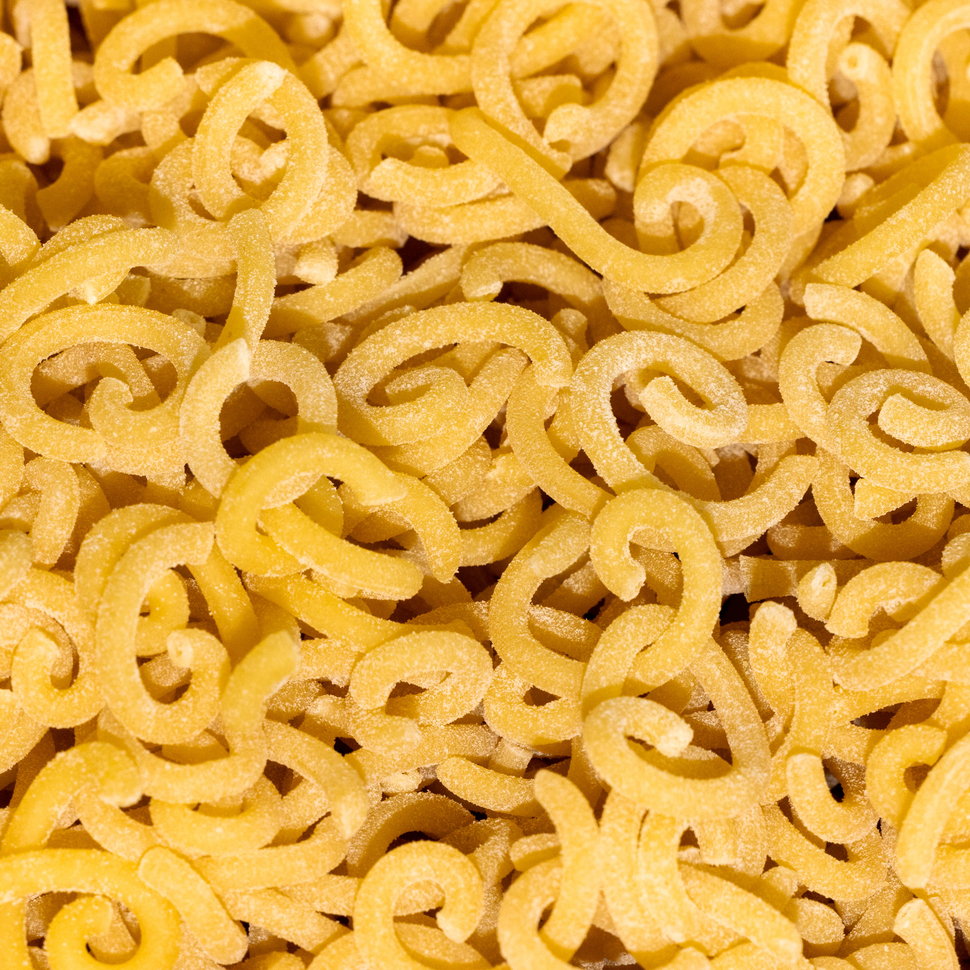 The image shows a close-up of uncooked, curly-shaped pasta, with a yellow-golden color and a lightly dusted surface. The pieces are irregularly twisted.