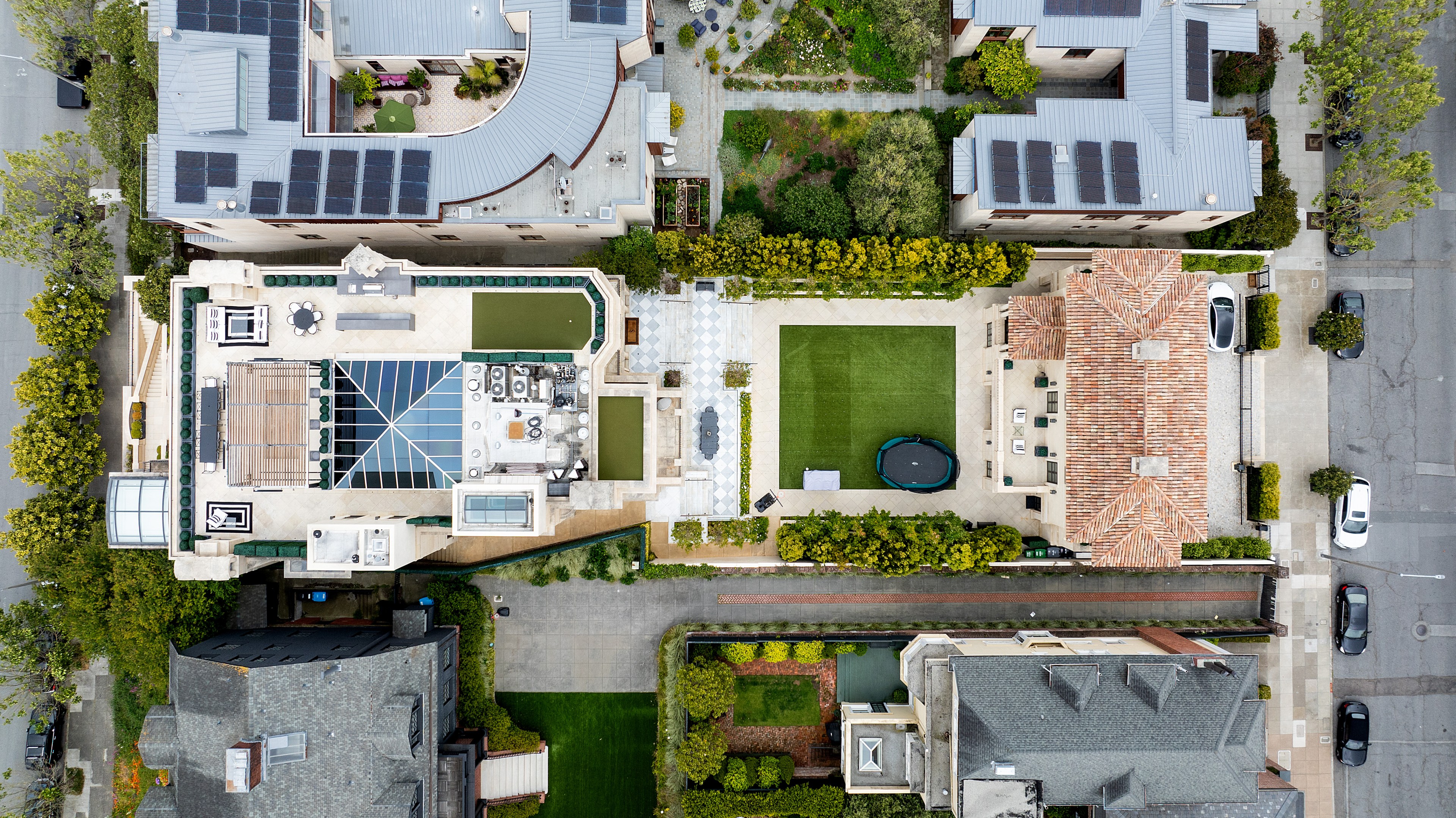 An aerial view shows a modern house with a rooftop patio, solar panels, outdoor furniture, and lawns. Surrounding homes and streets with parked cars are visible.