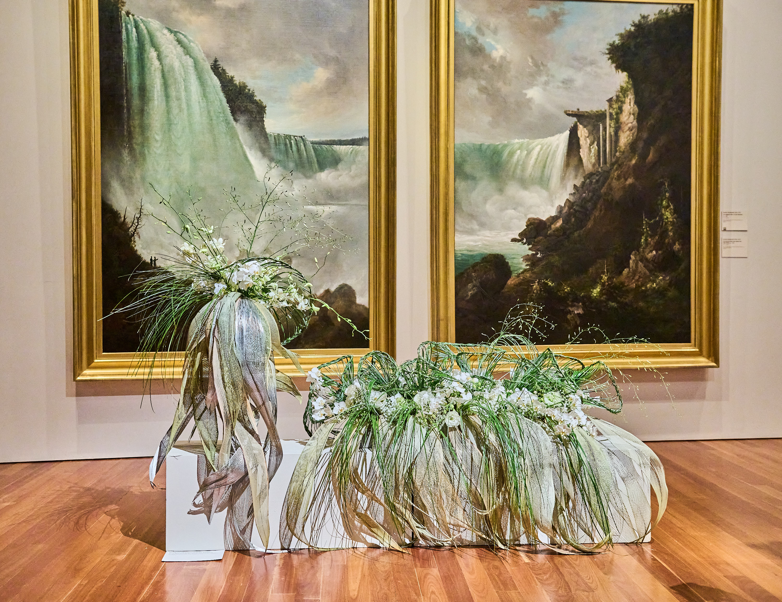 Two large paintings of waterfalls in gold frames are displayed on a wall. In front are two elaborate, contemporary floral arrangements on white pedestals.