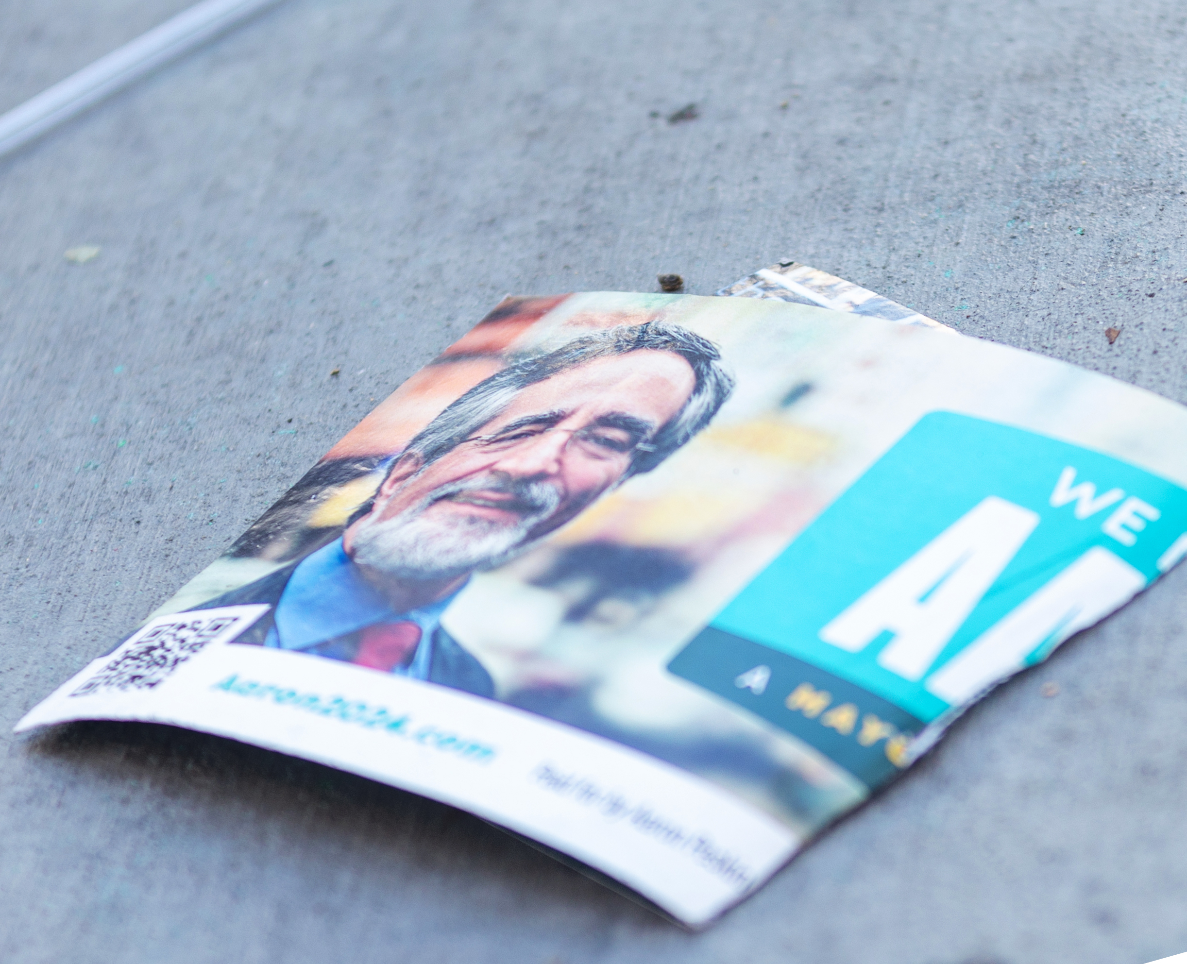 A torn political flyer on the ground shows Supervisor Aaron Peskin with a beard and suit. It includes a QR code, a website link, and partially visible text likely promoting a campaign.
