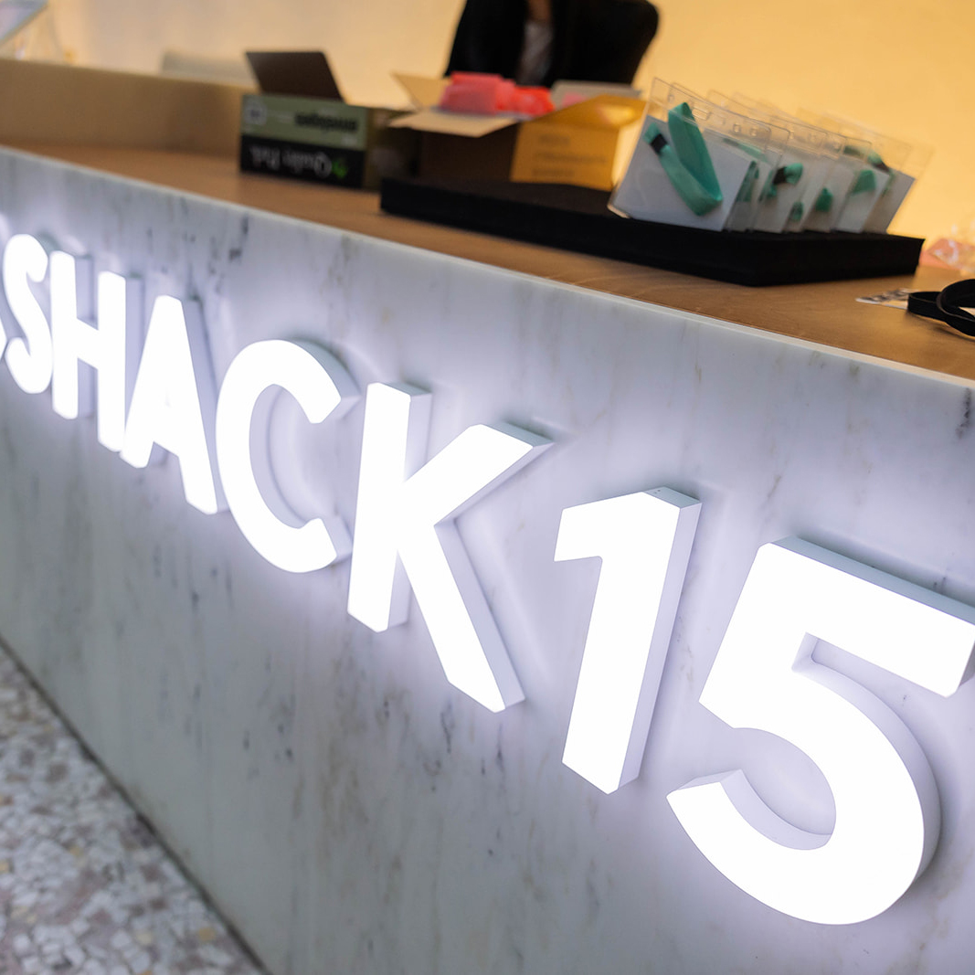 The image shows a reception desk with a person behind it and a sign with large, illuminated letters spelling &quot;SHACK15.&quot; There are also some boxes and items on the desk.