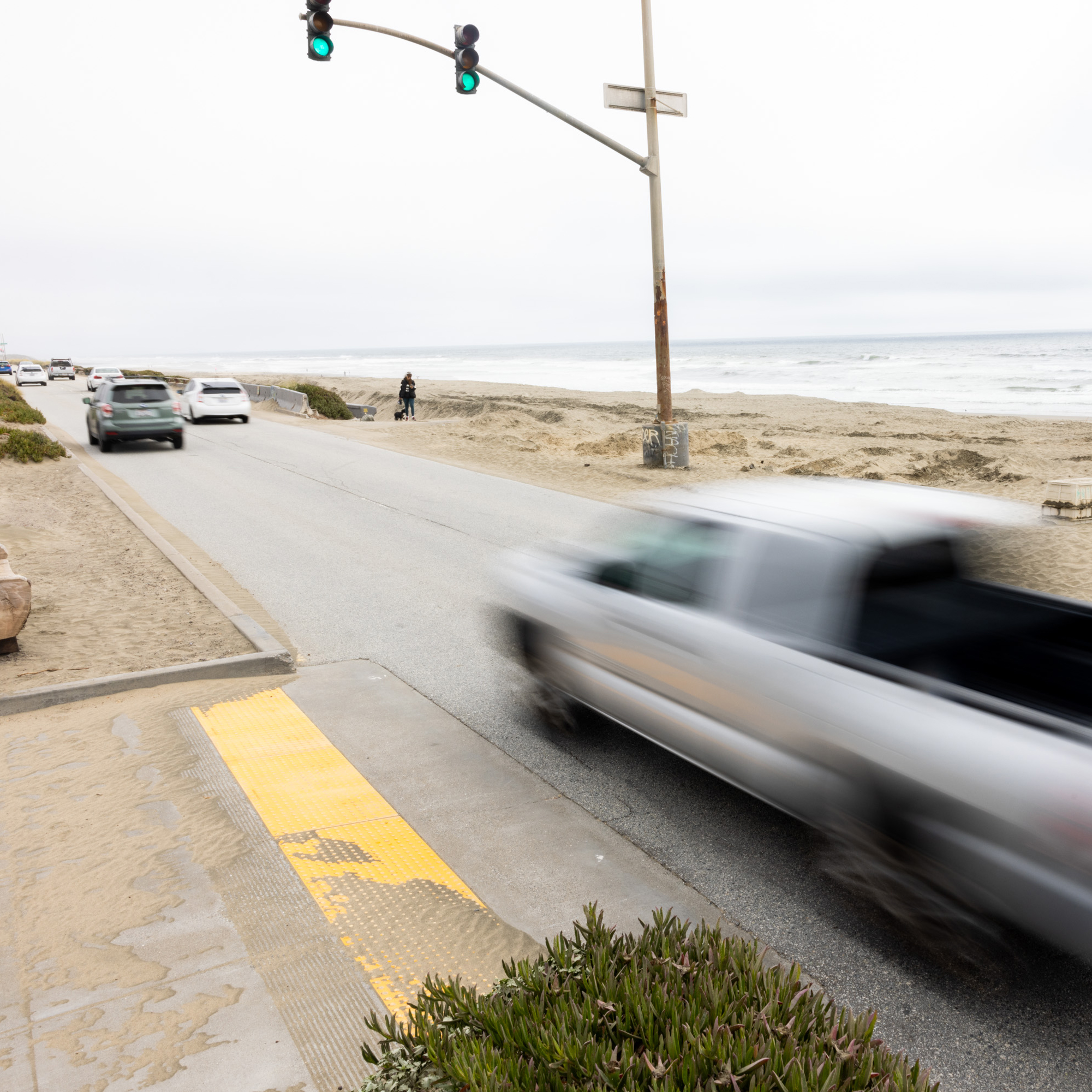 A road runs along a sandy beach with vehicles on it, including a speeding truck in the foreground. A person walks their dog near the ocean under a traffic signal.