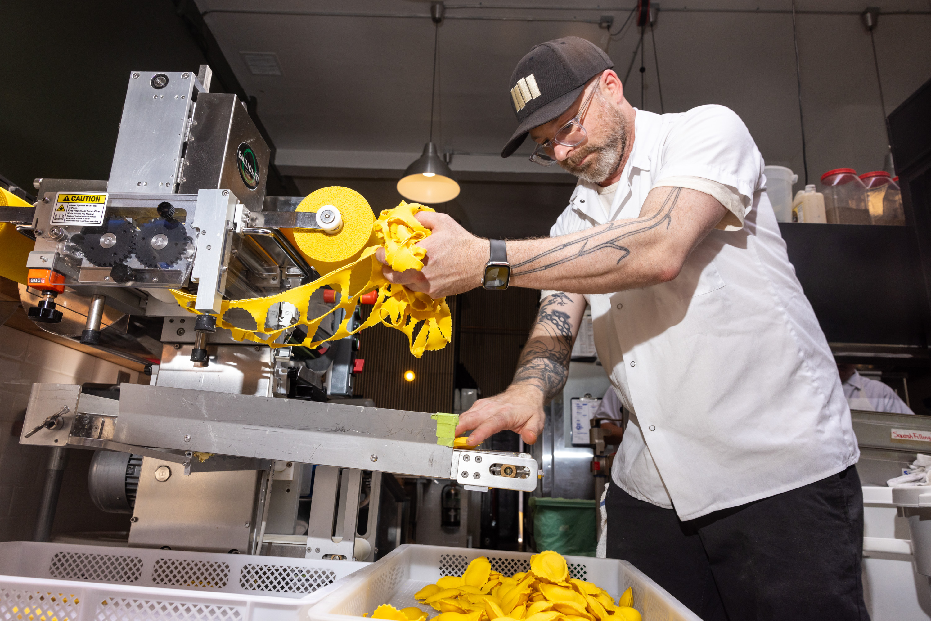 A man operates a pasta-making machine, feeding yellow pasta sheets into it. He's wearing a cap and glasses and is surrounded by bins containing yellow pasta.