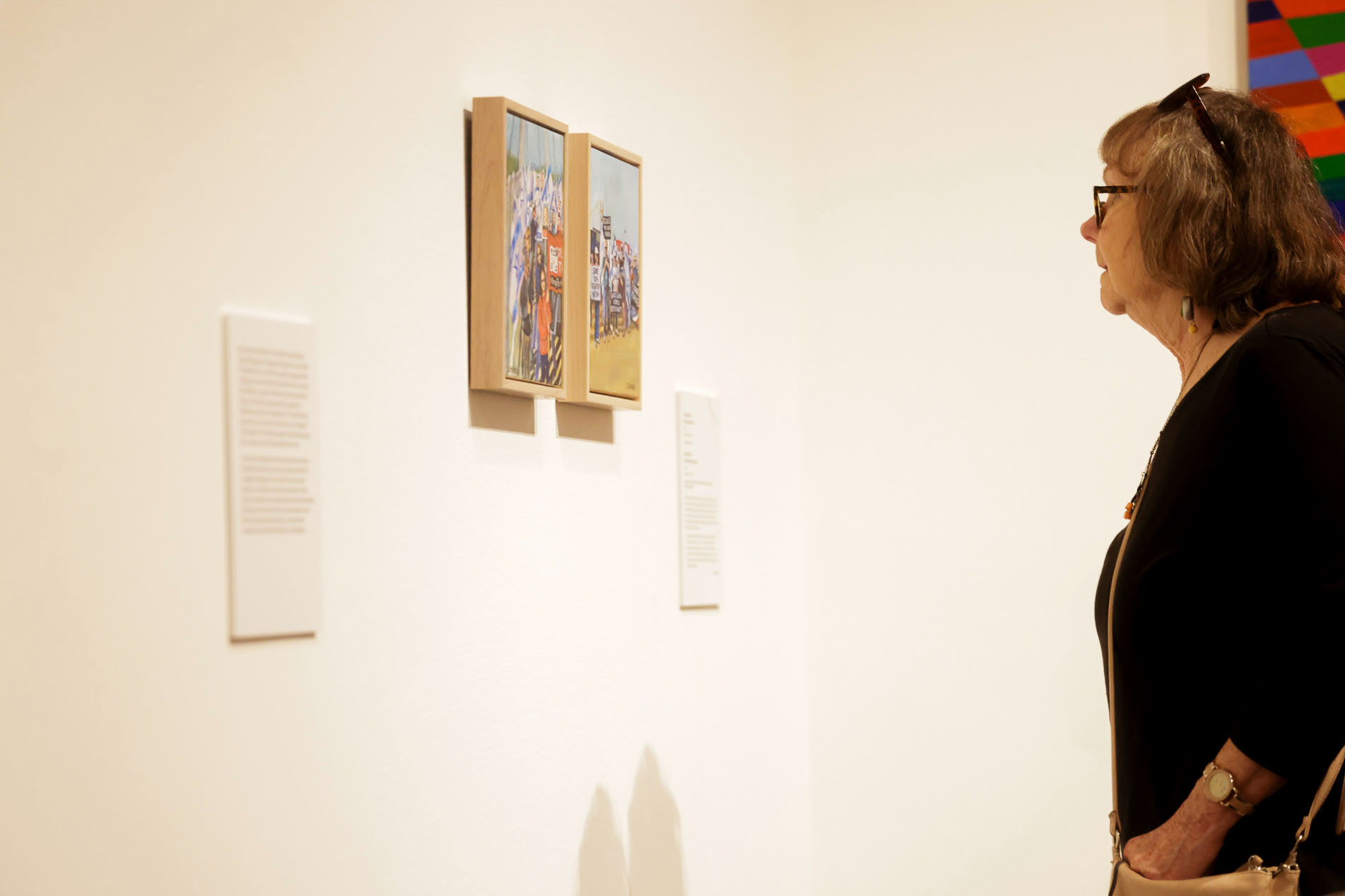 A woman with glasses and a handbag closely observes two small framed paintings in a gallery, with description placards on the wall beside them.