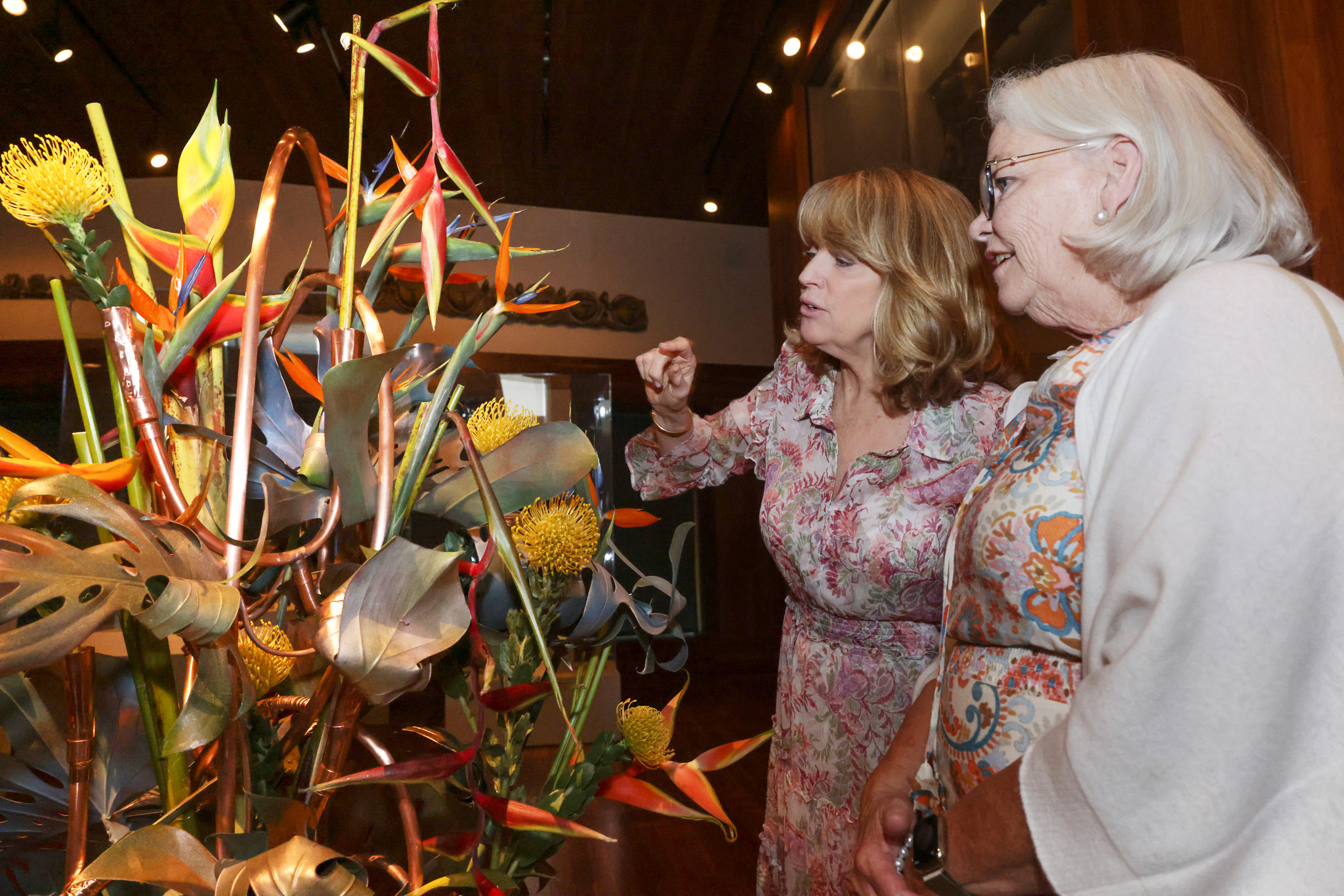 Two women, one elderly and one middle-aged, admire a vibrant floral arrangement featuring yellow and orange blooms in an indoor setting with soft lighting.