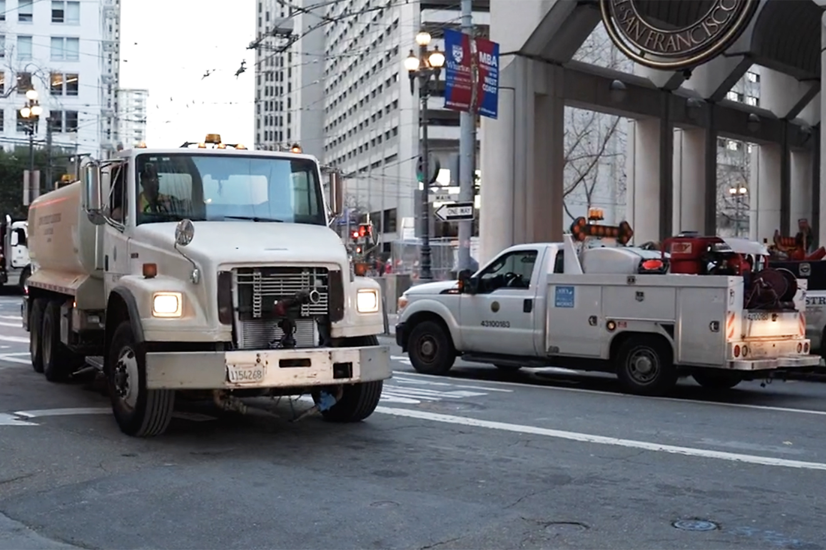 Two white service trucks are on a city street with tall buildings and streetlights.