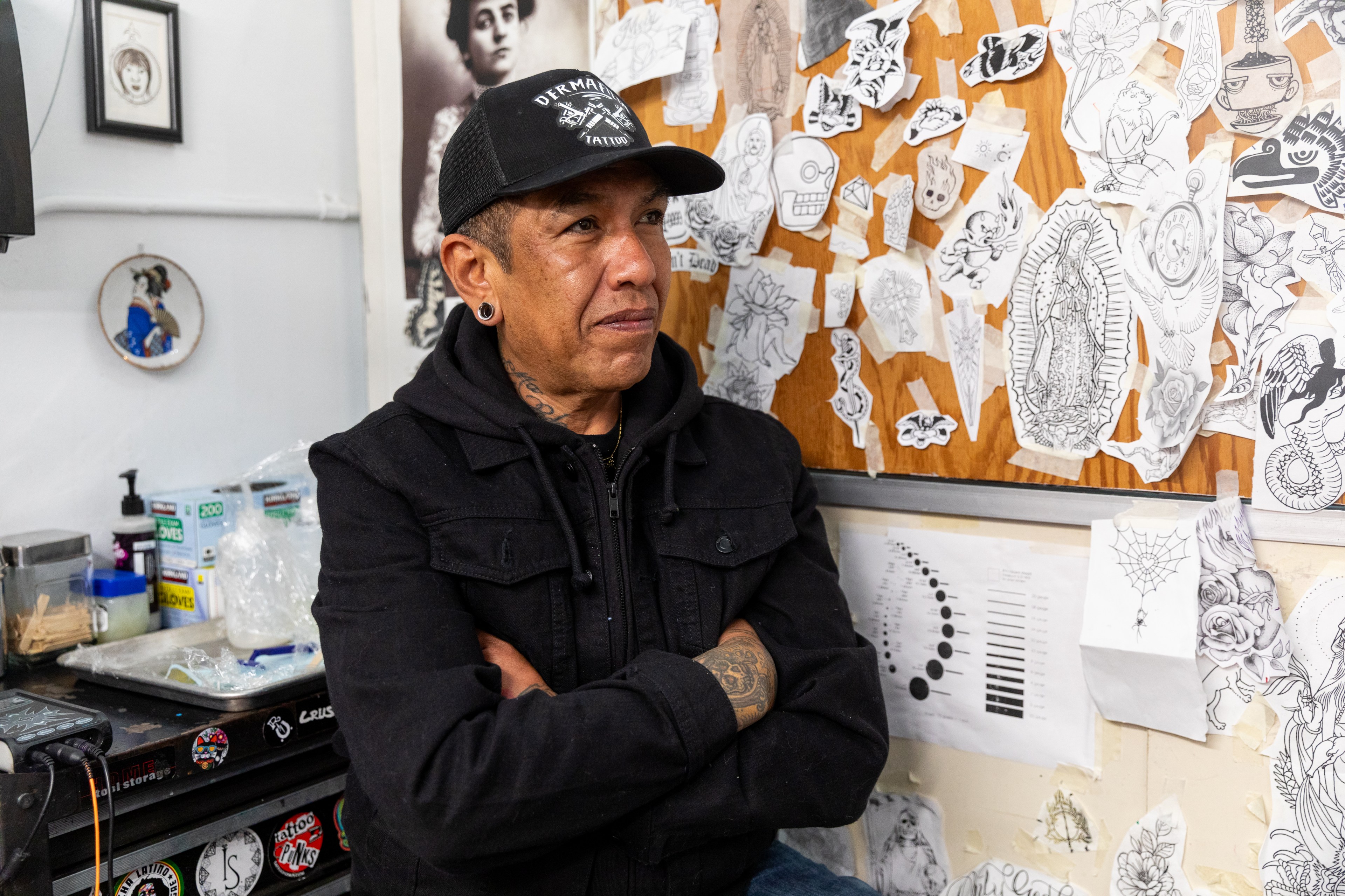 Roberto Barriga with tattoos, wearing a black jacket and cap, stands in a tattoo studio. The wall behind them is covered with various tattoo design sketches.