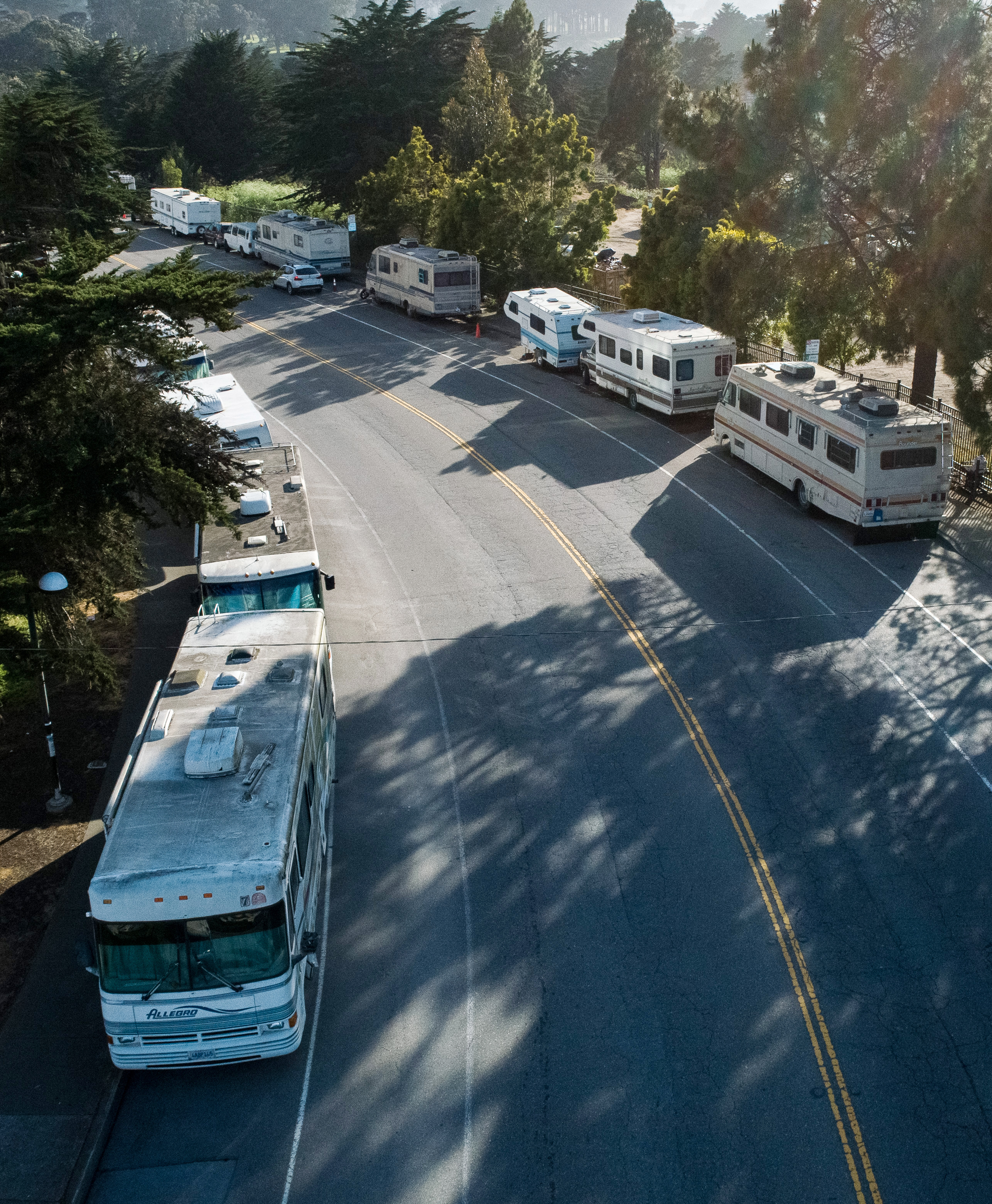 A curved street lined with multiple parked RVs on both sides, set against a backdrop of lush green trees and soft sunlight filtering through the branches.