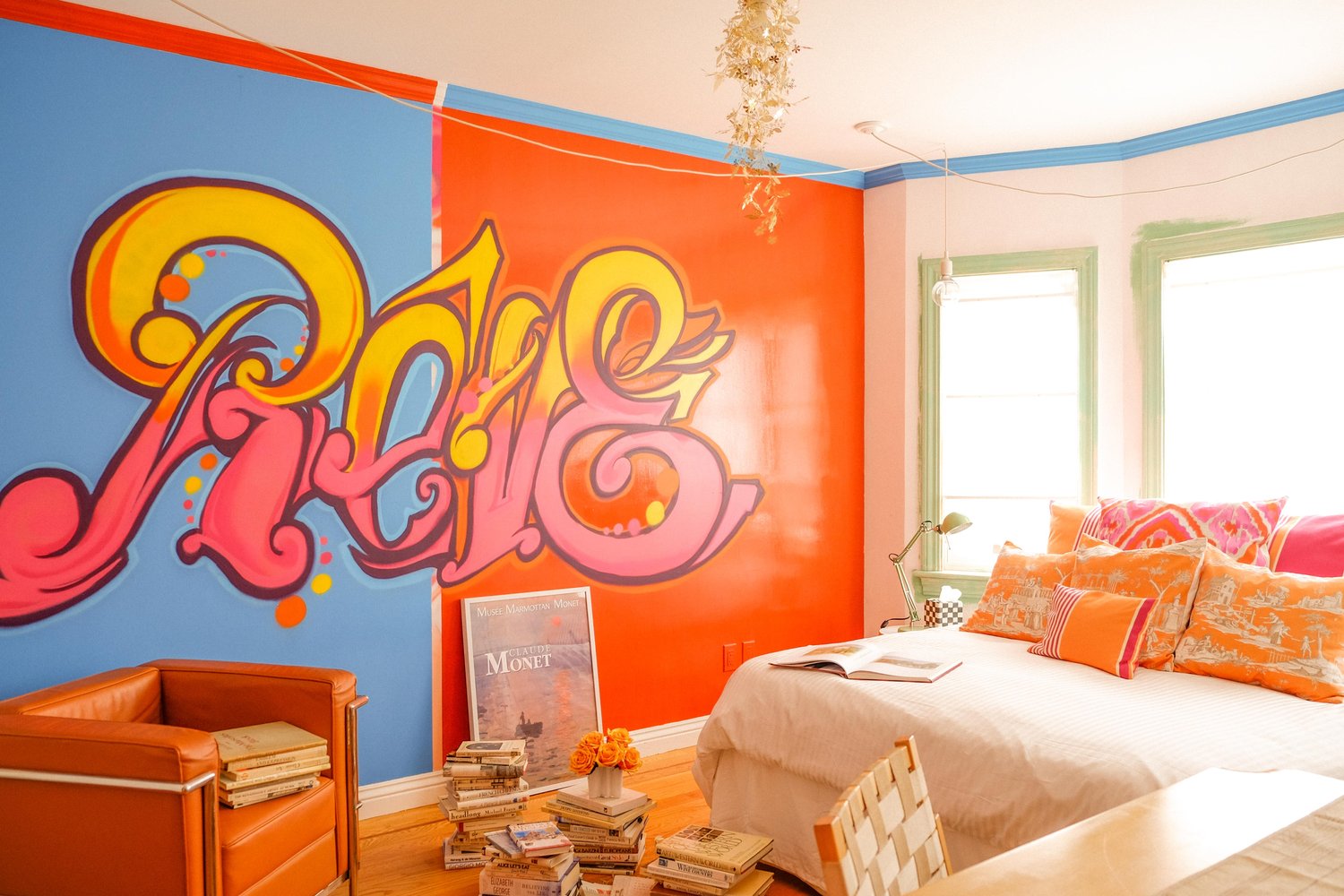 A vibrant room has an orange and blue wall with large graffiti art, an orange armchair, a bed with colorful pillows, stacks of books, and bright windows.