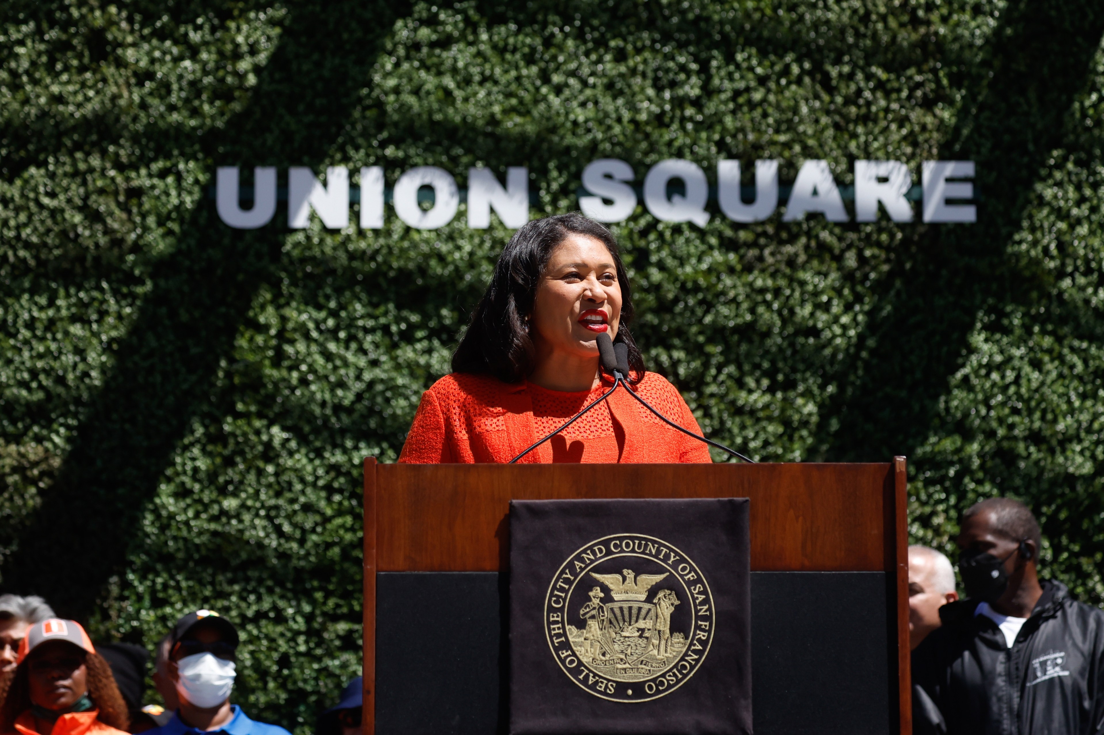A woman in an orange top is speaking at a podium with a San Francisco seal. Behind her is a green foliage wall with the words "Union Square" in large, white letters.