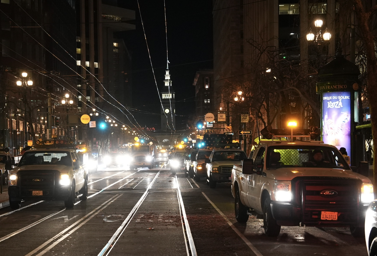 The image shows a night-time city street with multiple vehicles, mostly trucks, with headlights on.