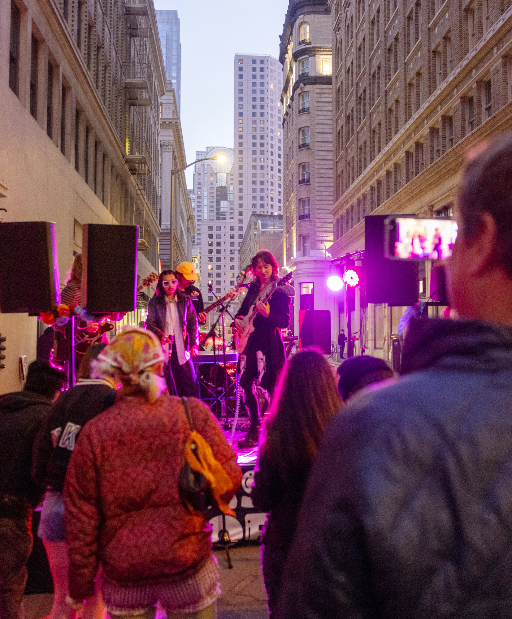 This image shows a street concert with four musicians performing, surrounded by tall buildings. Several people are watching, including someone filming with a smartphone.