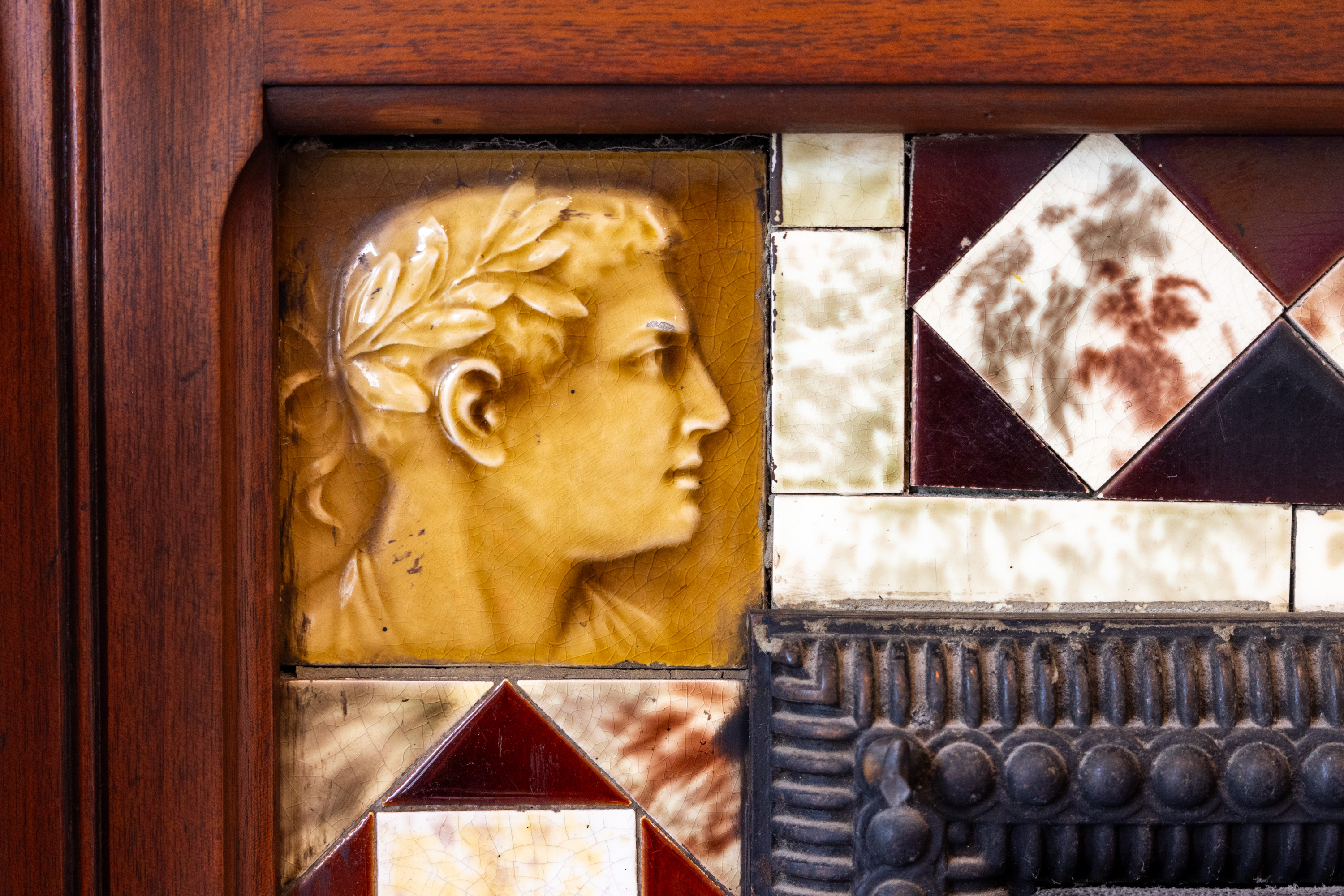 The image features a carved classical profile of a man's head with a laurel wreath, set in a wooden frame and surrounded by decorative tiles with various patterns.