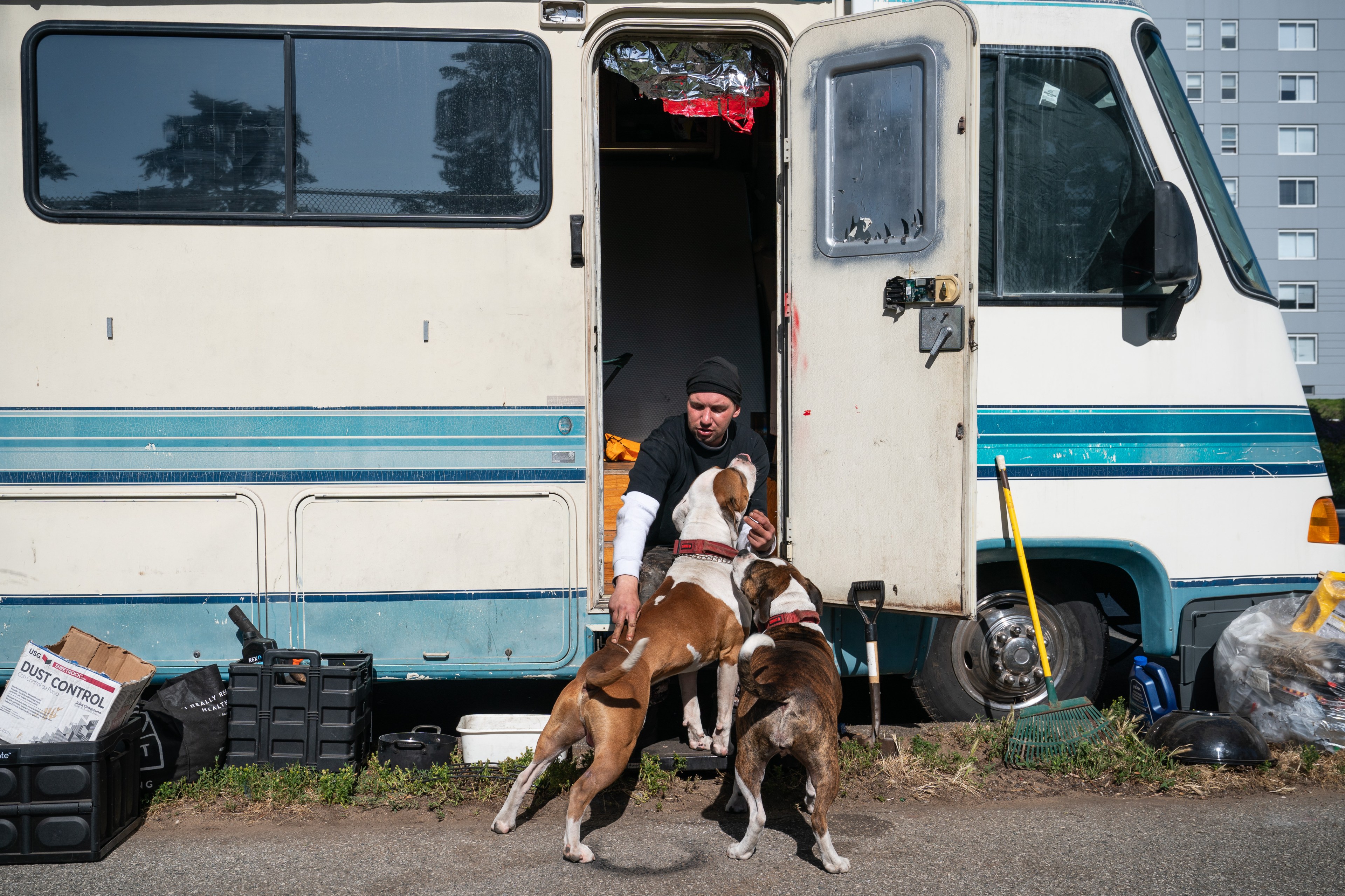 A person is crouching in the doorway of an RV, petting two eager dogs. The RV door is open, revealing some interior, and various items are scattered around outside.