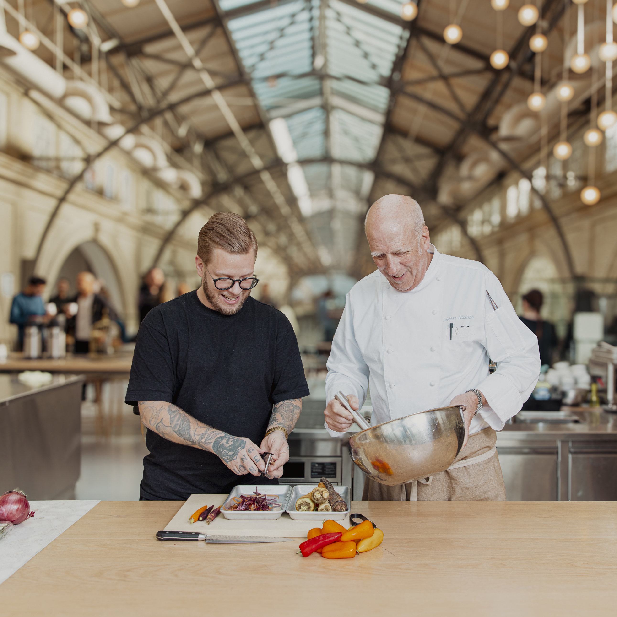 Two chefs, one in black and tattooed, the other in white, cook in a spacious, industrial-style kitchen. They're prepping vegetables and smiling together.