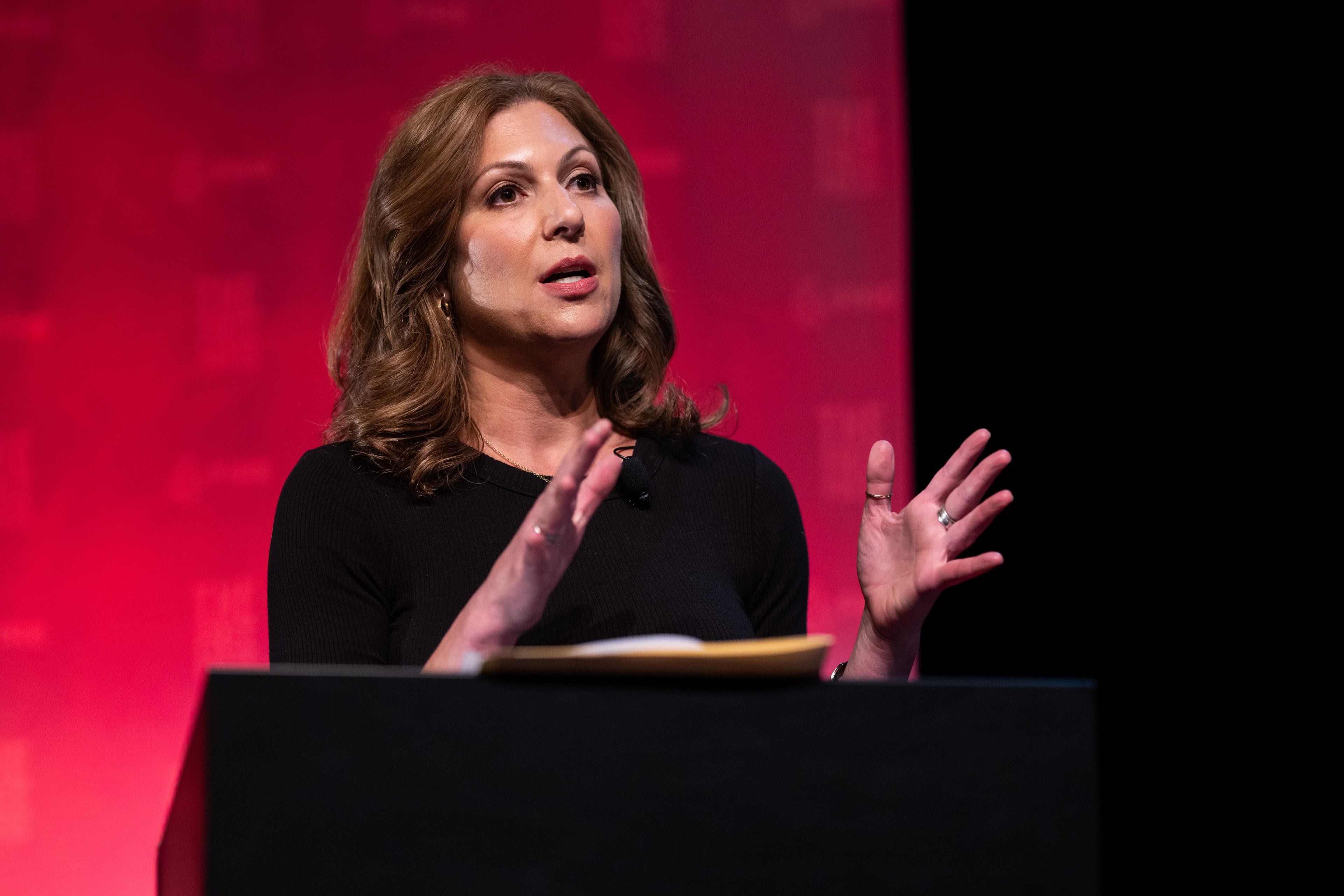 A woman with wavy brown hair is speaking passionately from behind a podium, gesturing with her hands. The background is red with a subtle pattern printed on it.